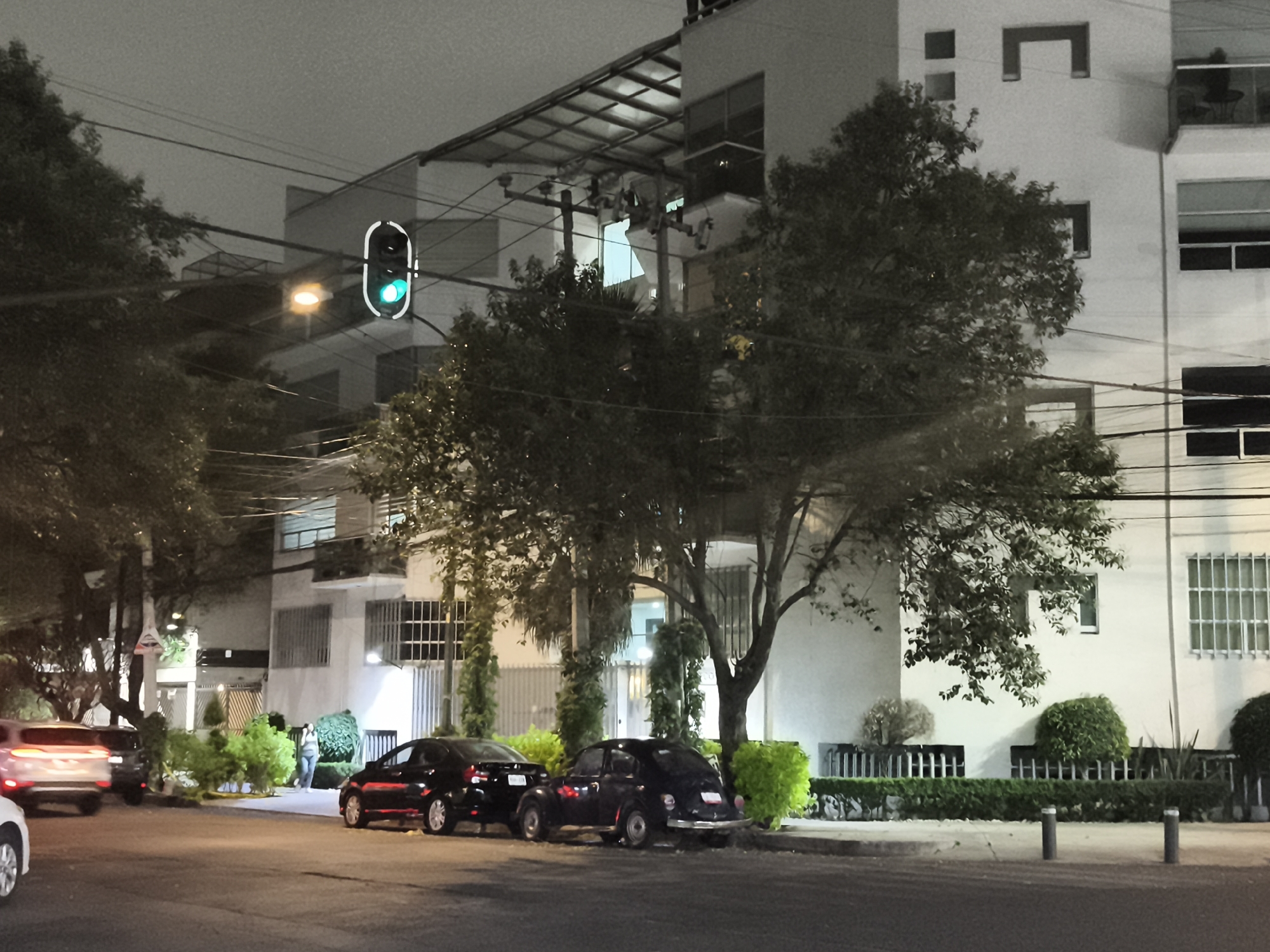 The image depicts a street scene at night in Mexico City, Mexico. A traffic light is visible with a green light illuminated. Two dark-colored cars are parked on the side of the road, including a Volkswagen Beetle. A white building with distinct architectural features, including a rooftop structure and window design, stands in the background, partially obscured by trees. A person is walking on the sidewalk. Additional cars and trees are visible in the distance. Power lines crisscross the sky.