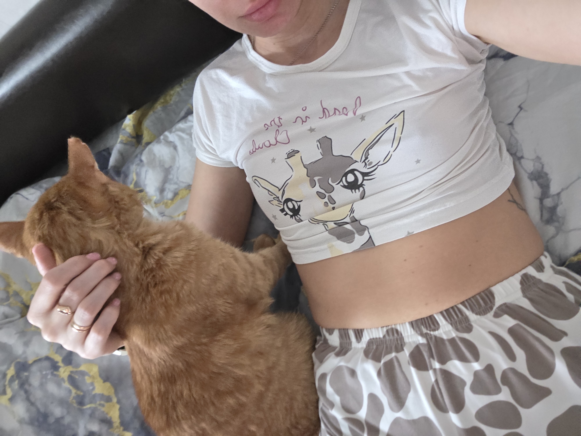 A woman lies on a bed with a gray and gold patterned comforter, holding an orange cat. The woman wears a white crop top with a giraffe graphic and brown and white spotted shorts. The cat is primarily in the lower left of the frame, being held by the woman's left hand. The location is Chaikovsky, Russia.