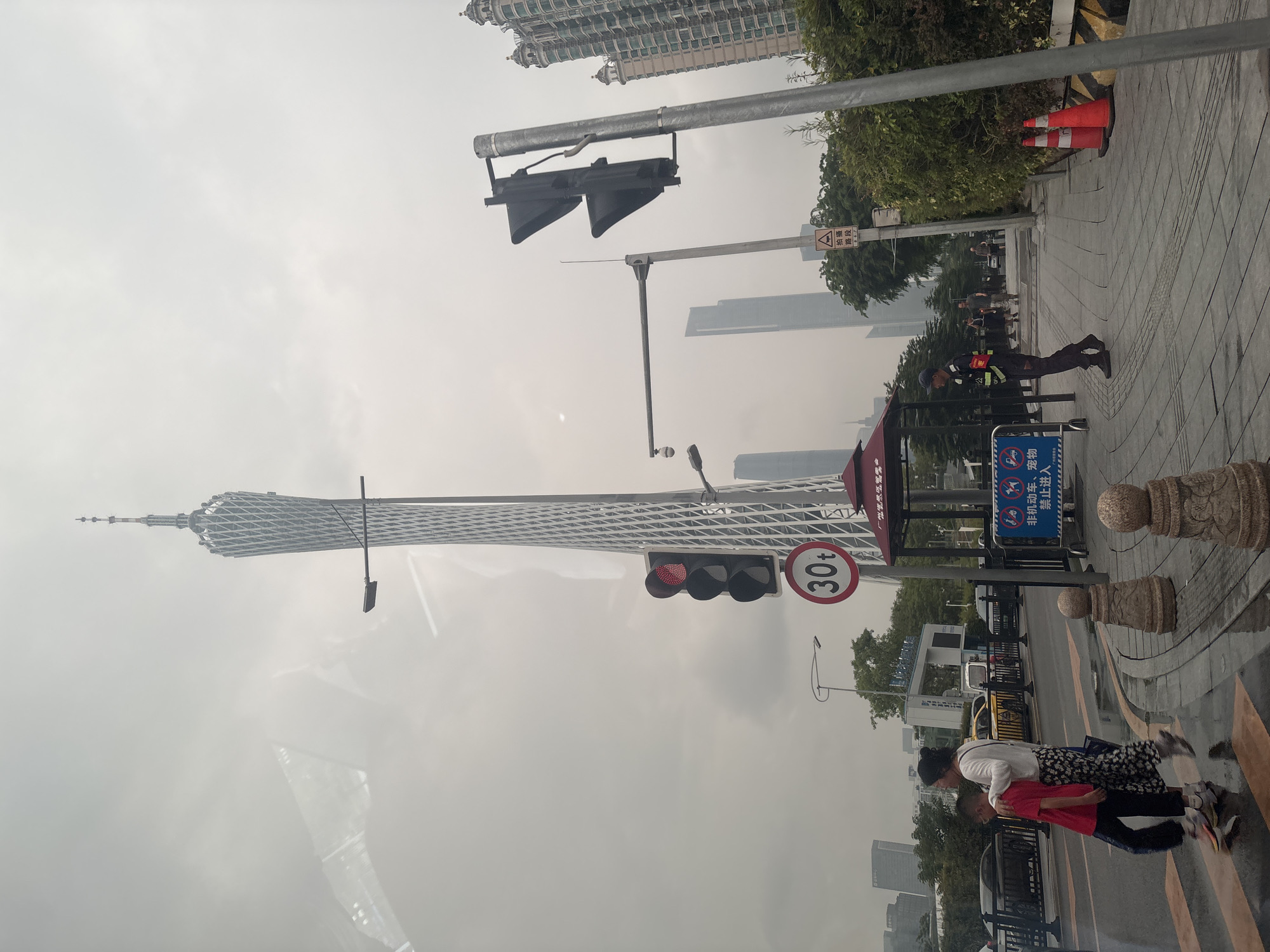 The image captures a street scene in Haizhu District, China, dominated by the Canton Tower in the background. In the foreground, a pedestrian crossing is visible with two individuals, a woman and a child, crossing from left to right. The woman is wearing a white long-sleeved shirt and a floral skirt, while the child is in a red t-shirt and dark pants. A traffic light is illuminated red, and a circular sign with "30t" is posted nearby. A security guard in uniform stands on the sidewalk to the right, facing the street. The sidewalk is paved with tiles, and decorative stone bollards are placed along its edge. In the mid-ground, other pedestrians are walking on the sidewalk, and vehicles are present on the road. Several tall buildings, including modern skyscrapers and an older, ornate building with multiple domes, are visible in the distance. The sky is overcast, creating a diffused light. The scene appears to be a typical urban environment with traffic management systems and pedestrian infrastructure.