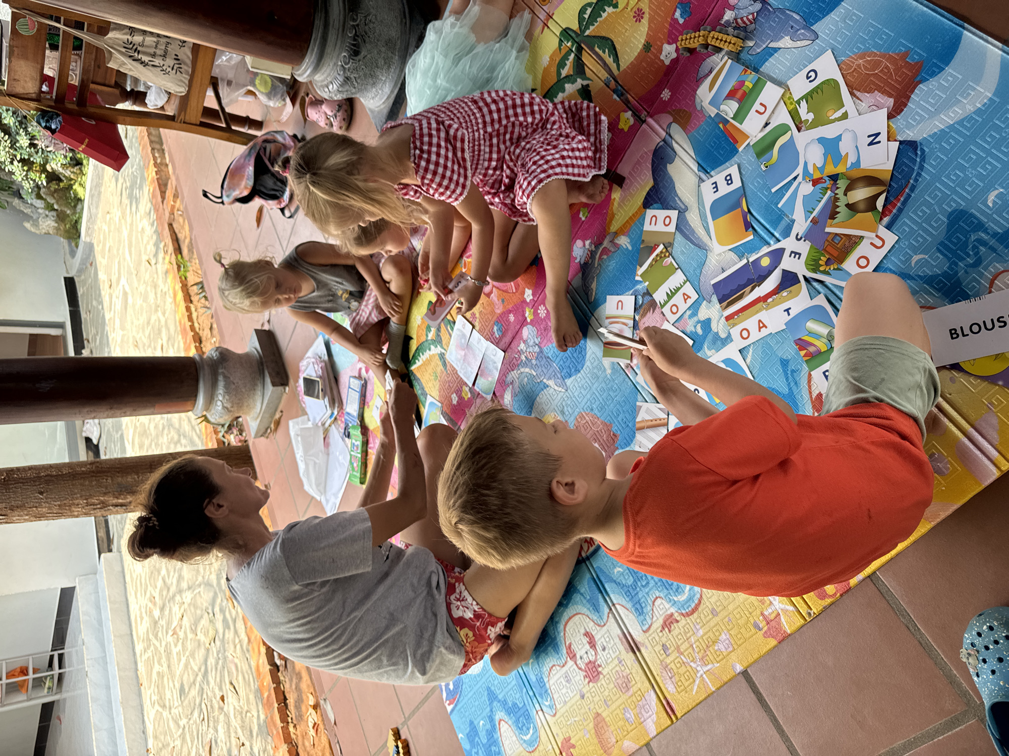 A group of an adult and several children are engaged in an educational activity on a colorful playmat laid over a tiled floor in what appears to be a covered outdoor area or patio in Mũi Né, Vietnam. The scene is brightly lit, suggesting daytime.
An adult woman, with her hair tied back, wears a grey t-shirt and sits on the left, looking down at the activity. To her right, a young boy in an orange t-shirt is intently focused on the cards. Next to him, a girl in a red and white checkered dress, and another girl in a grey top, are also interacting with the scattered cards. Several children are positioned around the mat.
The activity involves numerous rectangular cards, many displaying single letters such as 'N', 'O', 'U', 'A', 'E', 'B', 'G', 'Z', 'T', often accompanied by corresponding pictures. One white strip of paper near the boy's leg clearly shows the partial word "BLOUS," likely part of "BLOUSE."
The playmat features vibrant illustrations of dolphins, palm trees, and various patterns. The floor is made of terracotta-colored tiles, and large wooden columns are visible, supporting an overhead structure. In the background, beyond the pillars, greenery is visible, and items like a red box and a canvas tote bag, partially inscribed with "Shop local only good cherry," are seen. A blue Croc-style shoe is also visible on the far right. The atmosphere is one of focused learning and play.