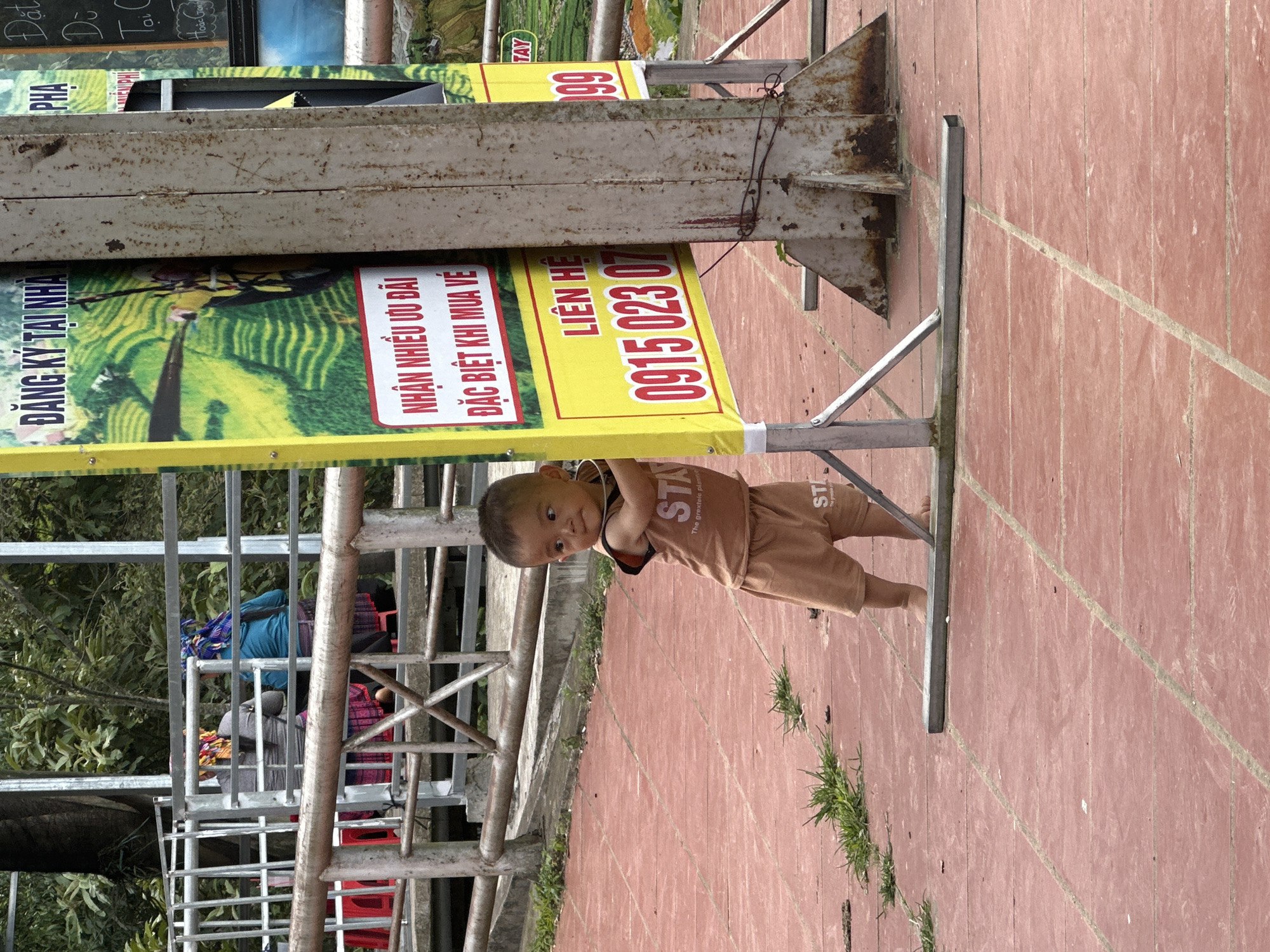 This image, captured in Tu Le, Vietnam, depicts a young child interacting with a large promotional sign in a public outdoor space. In the foreground, a toddler, appearing male and barefoot, stands on a reddish-brown paved surface characterized by light-colored grout lines forming a grid pattern. The child wears a light brown sleeveless top and matching shorts, both with partially visible text "STA" and "The grextele plann". He is holding onto the lower left vertical frame of a prominent yellow-bordered sign. The child's head is turned to his right, with a curious or slightly contemplative expression. Small patches of green grass are visible growing through cracks in the pavement, particularly to the left of the child. The sign the child is holding is large and yellow-bordered with a white interior, displaying Vietnamese text. The top portion reads "ĐĂNG KÝ TẠI NHÀ" (Register at home) and features an image of green terraced rice fields. Below, a red banner contains the text "NHẬN NHIỀU ƯU ĐÃI ĐẶC BIỆT KHI MUA VÉ" (Receive many special offers when buying tickets). Further down, "LIÊN HỆ" (Contact) is displayed above a red phone number beginning with "0915 023 0". The sign is supported by a silver-colored metal frame forming a triangular base on the ground. To the right of this sign, a heavily rusted, light brown-grey vertical metal pole partially obscures a similar yellow-bordered sign, from which only a partial red number "99" is visible. This pole is also supported by a large, triangular metal base. In the mid-ground, behind the child and signs, a silver-colored metal railing or fence structure is present, consisting of horizontal and vertical bars. A section of a darker, possibly rusty, handrail is visible in front of this structure. Behind the railing, two individuals, likely women, are seated with their backs to the viewer. They are wearing distinctively patterned head coverings – one with yellow, orange, and red patterns, and the other with blue and purple patterns. The person on the left wears a grey long-sleeved top and a patterned skirt, while the person on the right wears a blue long-sleeved top and a darker patterned skirt. They appear to be sitting on red plastic stools or benches. The background consists of dense green foliage and trees, indicative of a natural, possibly mountainous or hilly, environment. A partial black chalkboard sign with white text, including "Hoàn G...", is visible on the