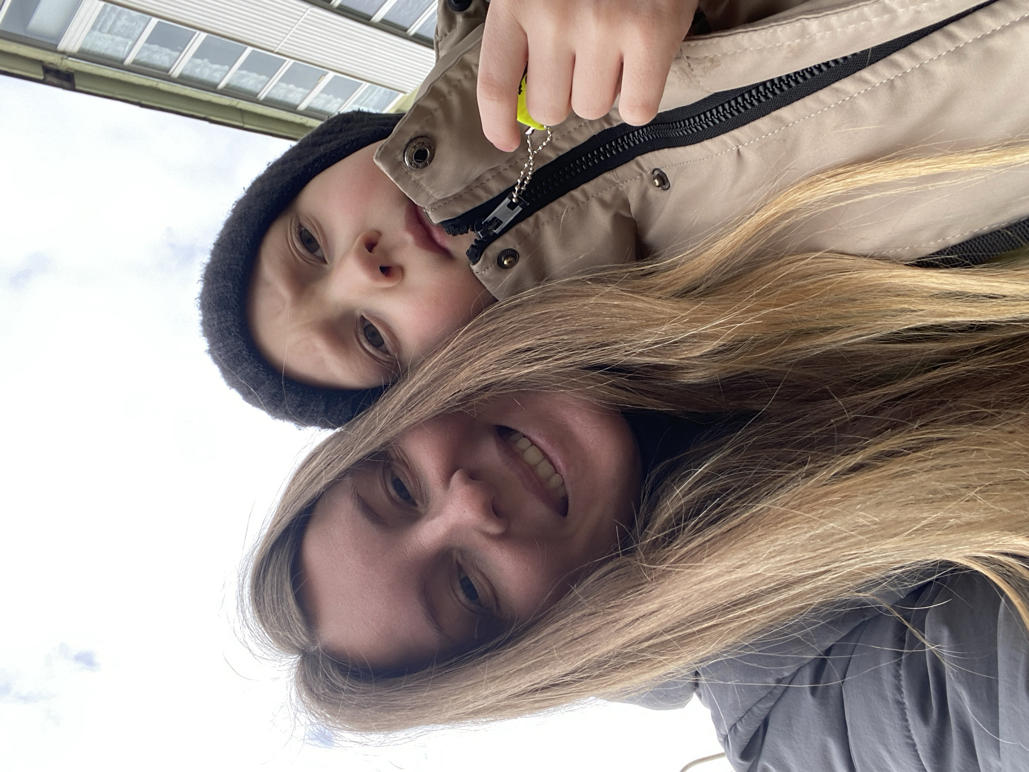 A low-angle, close-up photograph shows two individuals, likely a woman and a child, posing for a selfie. The woman is positioned in the foreground, smiling slightly, with long, light brown hair partially obscuring her face. She is wearing a gray jacket. Behind her is a child wearing a dark beanie and a tan winter coat. The child has fair skin and is looking at the camera. Their right hand is visible and they are holding a neon yellow object attached to a silver chain. The background shows a light sky with a building visible. This photo was most likely taken in the city of Noviy, Russia.