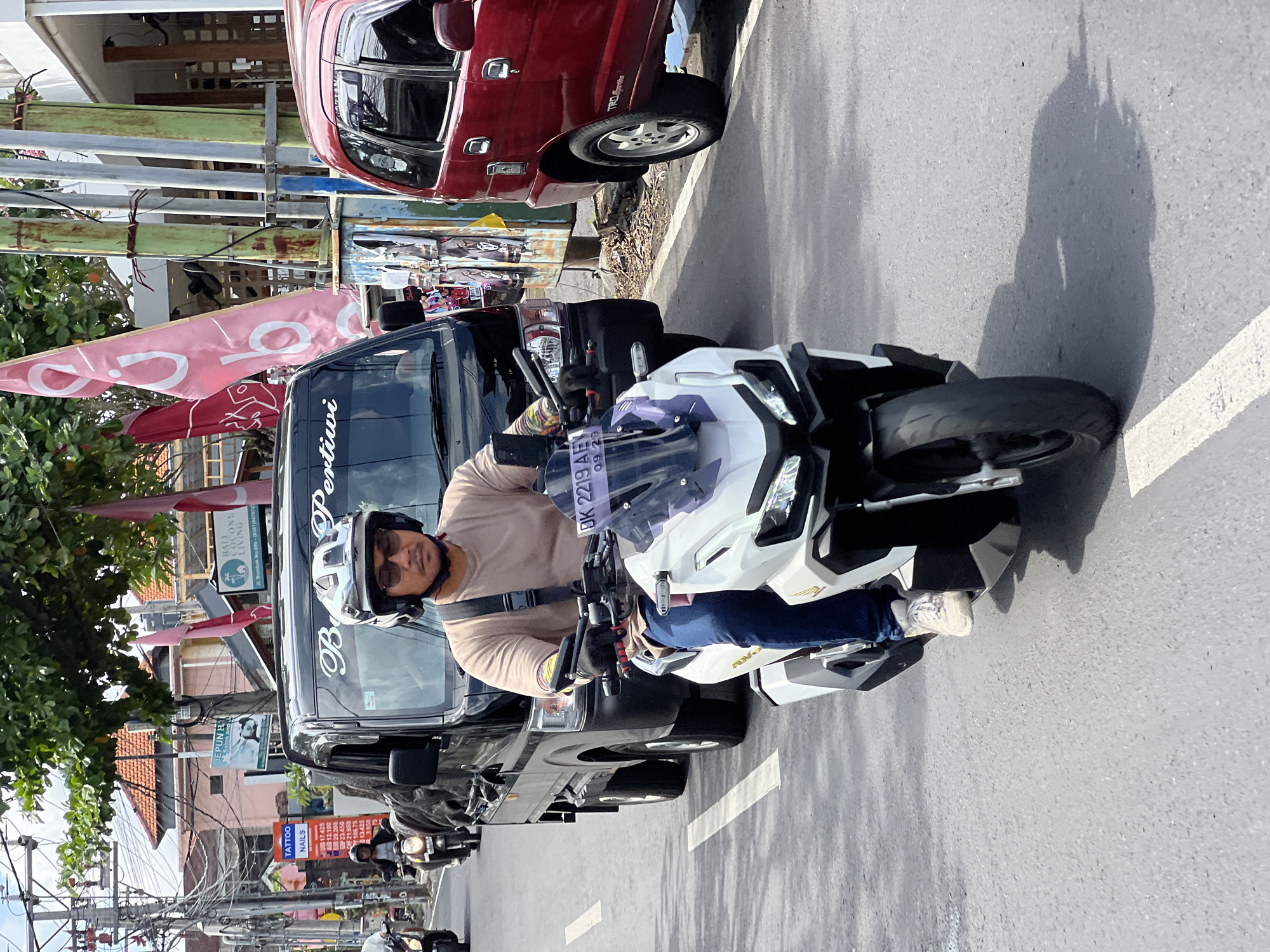 This is a medium shot, taken from the right side of the road, of a man riding a white scooter on a sunny day in North Kuta, Indonesia. The man, wearing a beige t-shirt, blue jeans, white sneakers, and a grey helmet with dark sunglasses, is looking forward. He is riding in the middle of the road, on the right side of a black truck, which is in front of a red SUV. The scooter has a white body with black accents and a clear windshield. The license plate on the scooter is visible and reads "DK 2219 ABY". The road is paved with white lane markings. In the background, there are buildings and trees, with power lines visible overhead. Several flags are also present, including two large pink flags with white text. The scene appears to be a typical street scene in a busy Indonesian city.
