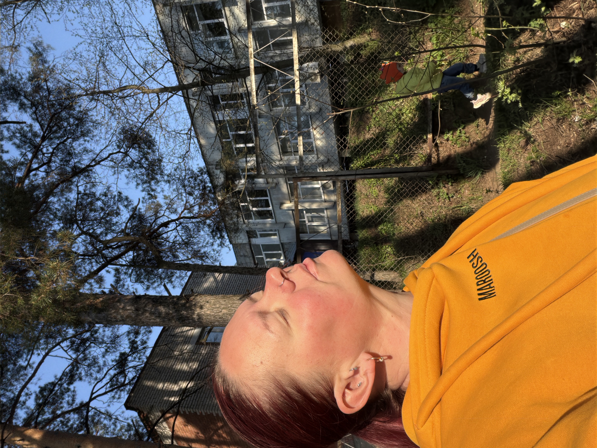The image shows a person with fair skin and reddish-brown hair, wearing a bright yellow hoodie with the word "MAROOSH" printed on it. They are looking upwards with their eyes closed. The background includes trees, a light-colored building with multiple windows, and a chain-link fence. A person in a light-green jacket and blue pants is visible behind the fence. Based on visual cues, the location is in Chaikovsky, Russia.