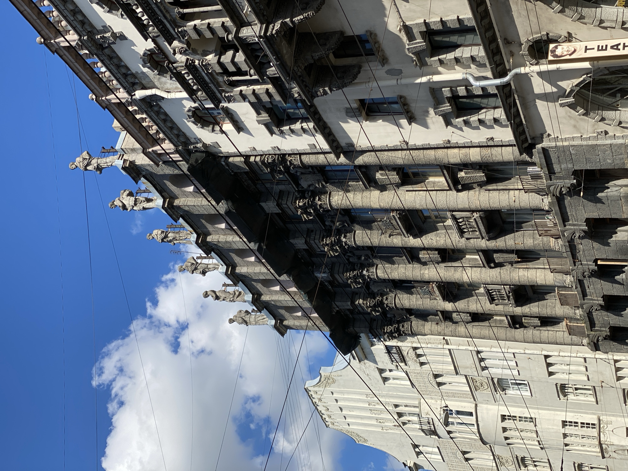 This media file captures an upward-angled view of two ornate buildings in Saint Petersburg, Russia, against a bright blue sky with scattered white clouds. The original image is oriented vertically, showing the buildings stretching upwards towards the sky.
The prominent building on the left and center is a striking, dark grey structure characterized by its elaborate architectural details. Multiple large, sculpted figures, possibly atlantes or caryatids, protrude from the facade at various levels, adding a distinctive decorative element. Below these figures, the building features numerous fluted columns, intricately framed windows, and textured stonework that creates strong shadows in the bright sunlight. Dark roofing elements are visible at the very top. Many thin overhead utility wires crisscross the scene, passing in front of the buildings and against the sky.
To the right, a second building with a lighter, creamy white facade is partially visible. It displays simpler yet decorative architectural details, including textured panels and cornices, complementing the grandeur of its neighbor.
The scene depicts a clear, sunny day, likely morning or afternoon, given the sharp shadows cast by the architectural features. On the upper right section of the darker building, a vertical sign is visible, featuring a smiling woman's headshot and the text "ТЕАТР" (TEATR), which is Russian for "Theater." A grey metal pipe also runs horizontally across an upper section of the facade. The overall impression is one of majestic, historic urban architecture.