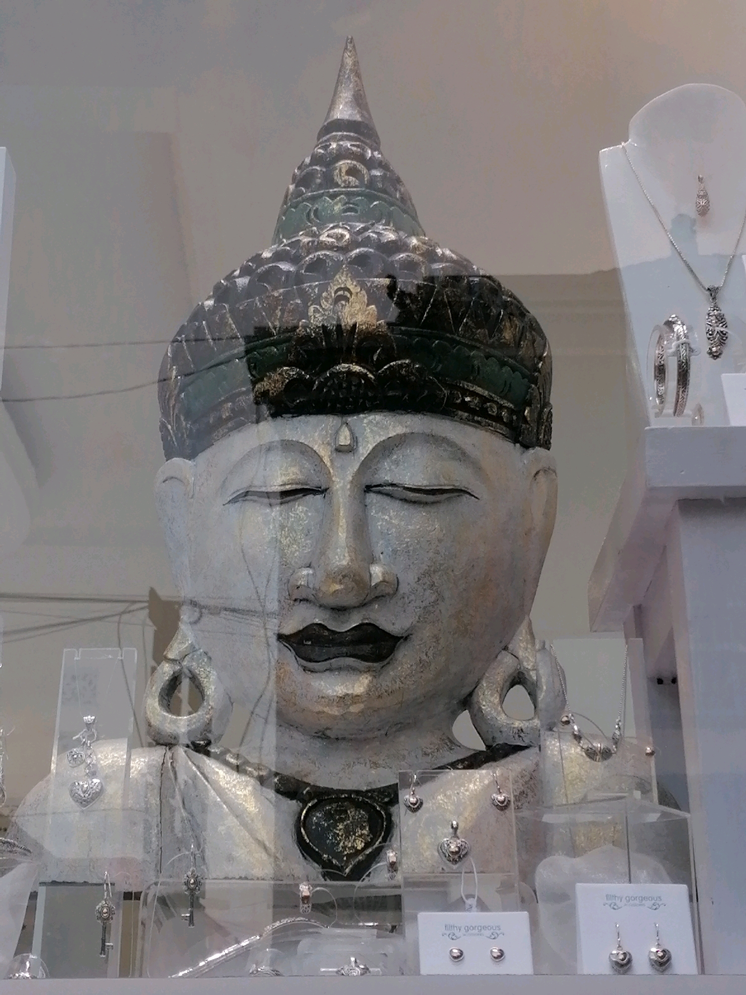 The image shows a display of jewelry in a shop window, with a large, ornate statue of a Buddha's head in the foreground. The statue is predominantly white with gold and dark green accents, featuring closed eyes and serene facial expression. It is adorned with a tall, pointed headdress and large earrings. Surrounding the statue are various jewelry pieces, including necklaces, earrings, and pendants, displayed on white stands and in clear plastic cases. Some of the jewelry features heart shapes and intricate designs. The backdrop appears to be a simple, light-colored wall. The overall scene suggests a retail environment, likely a jewelry store. The location is identified as Ubud District, Indonesia.