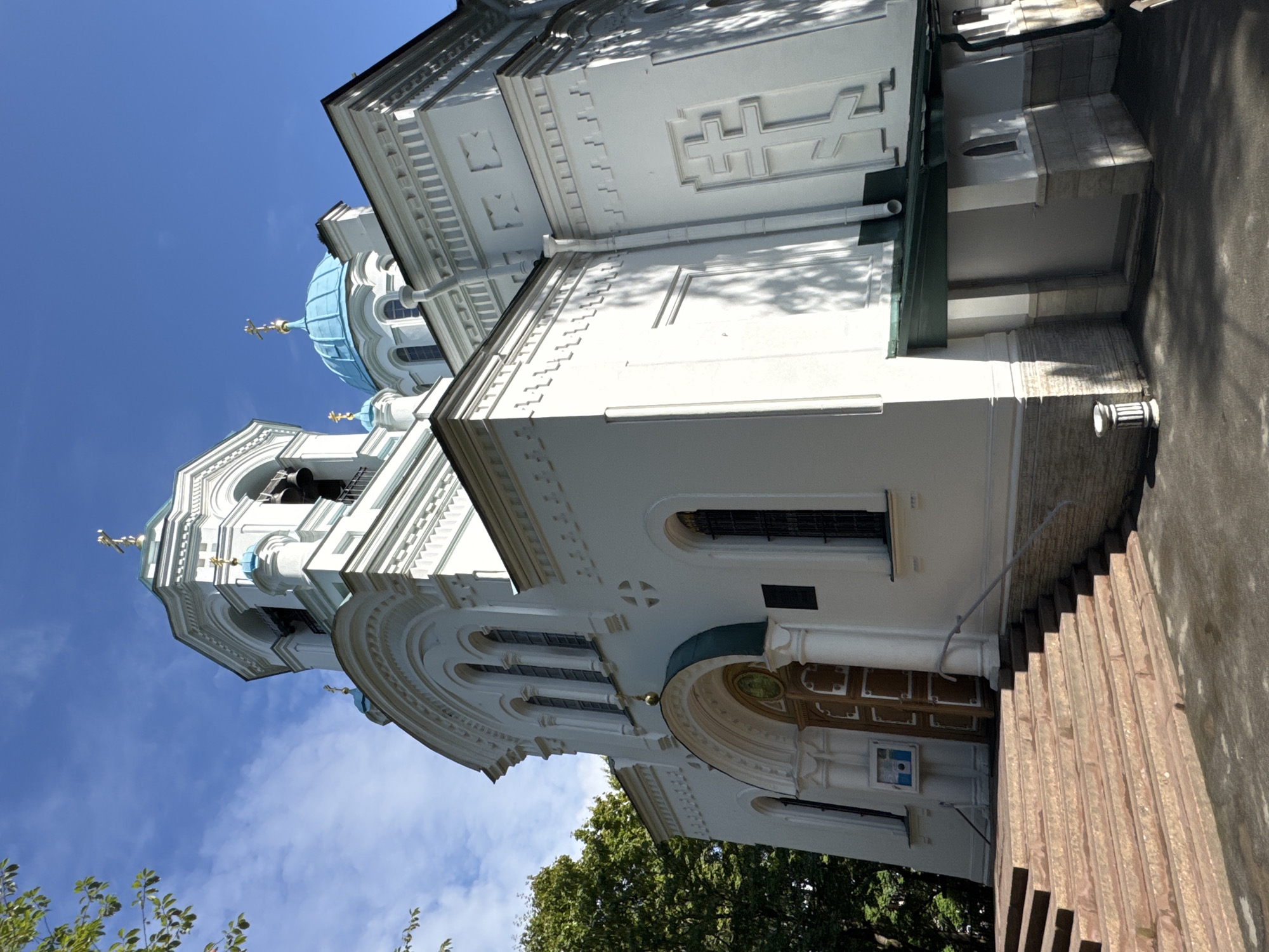 This is a low-angle, full exterior shot of the Church of the Assumption of the Most Holy Theotokos (Assumption Cathedral) located in Saint Petersburg, Russia, on the grounds of the Smolny Institute. The building is a white, neo-Byzantine structure with blue domes and a bell tower. Sunlight casts shadows on the building and the surrounding paved area, which is partially covered by dappled shade from trees to the left and right. A set of wide, stone stairs leads up to the main entrance. The sky is blue with scattered white clouds. The scene is devoid of people or any activity.