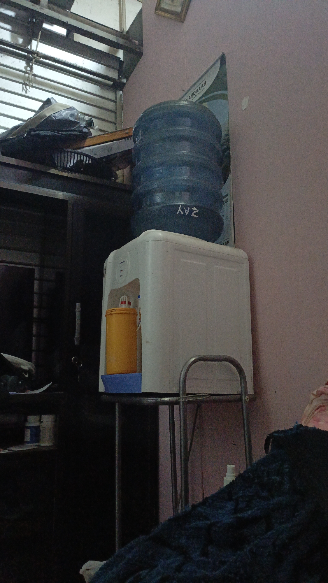The image captures an indoor scene, likely a room, with a water dispenser prominently featured in the center. A large, blue water bottle sits atop the white dispenser. To the left of the dispenser, a dark cabinet is visible, with some items on shelves above it, including what appears to be a black bag and a white and blue container. A yellow dispenser is positioned in front of the water dispenser. The background consists of a pinkish wall with a poster or banner hanging on it. The lighting is dim, suggesting it might be evening or the room is not well-lit. The location is identified as Gusunge, Indonesia. The scene appears to be domestic, possibly a home or a small office space. There are no people visible in the image.