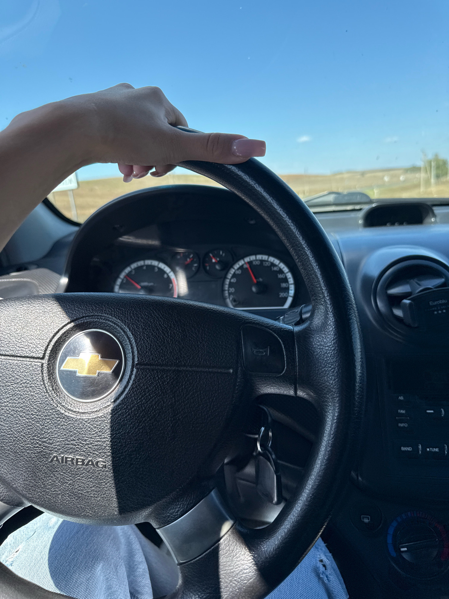 A first-person perspective shot shows a driver's hand with pink-painted fingernails resting on the steering wheel of a car. The hand is positioned at the top of the steering wheel, which features a Chevrolet emblem in the center and the word "AIRBAG" below it. The dashboard shows a speedometer with the needle indicating a speed of approximately 60 km/h. The interior of the car is visible, including parts of the dashboard, air vents, and a car key attached to the steering column. Outside the car, a bright blue sky and a distant, flat, grassy landscape suggest an outdoor, possibly rural setting. The car appears to be in motion. The provided information states the location is Iordanovca, Moldova, which is a village, not a city.