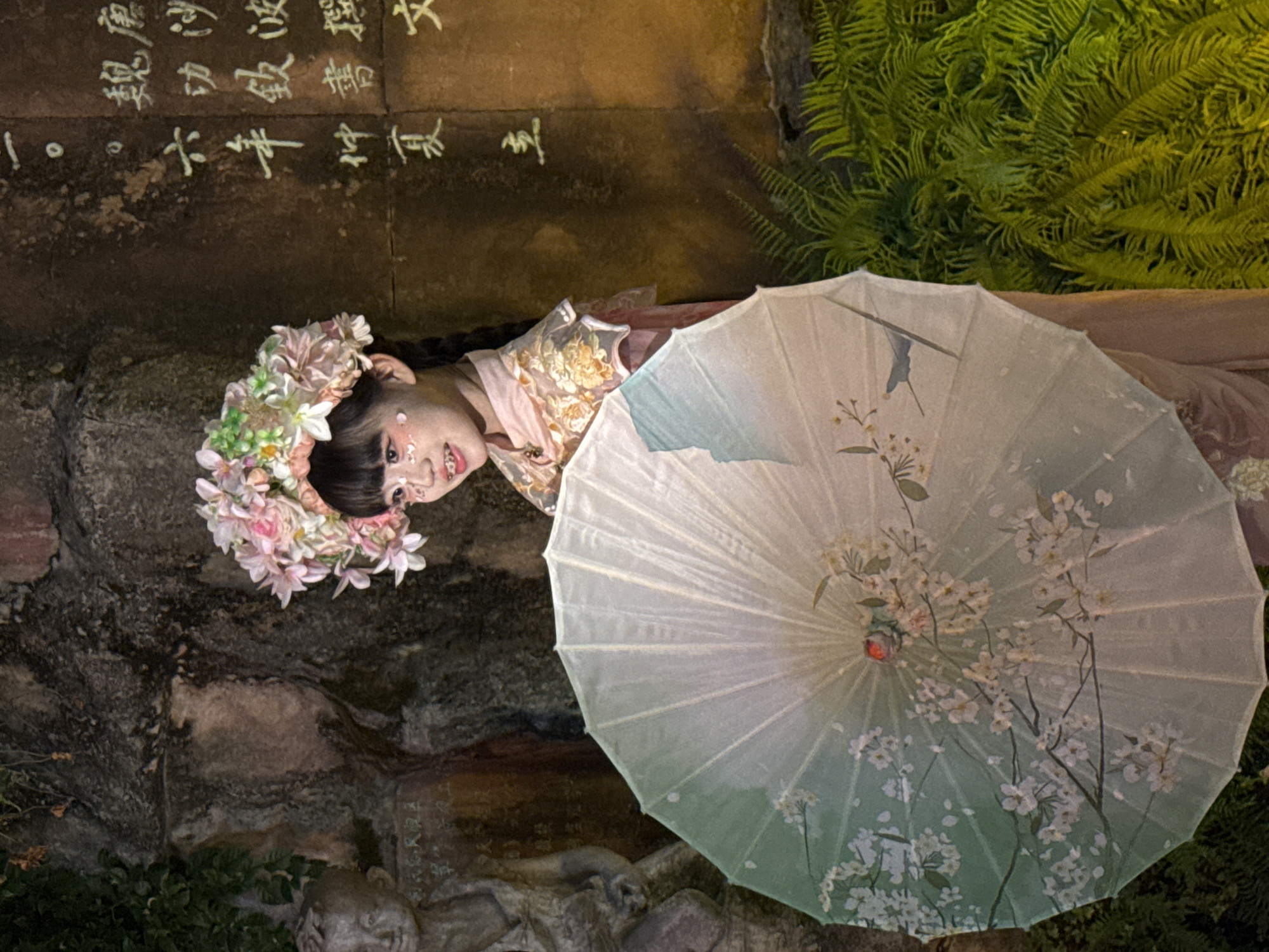 A young girl is standing in front of a stone wall with Chinese calligraphy, holding a decorative umbrella. The girl is wearing a floral headdress and a pink and gold outfit. Her face is adorned with makeup, and she has braces on her teeth. The umbrella is light green with a cherry blossom design. To the left of the girl, there is a stone statue of a child. The background also shows lush green foliage. The scene appears to be an outdoor setting, possibly in Yuzhong District, China, given the cultural elements present. The lighting suggests it is nighttime or a dimly lit area.