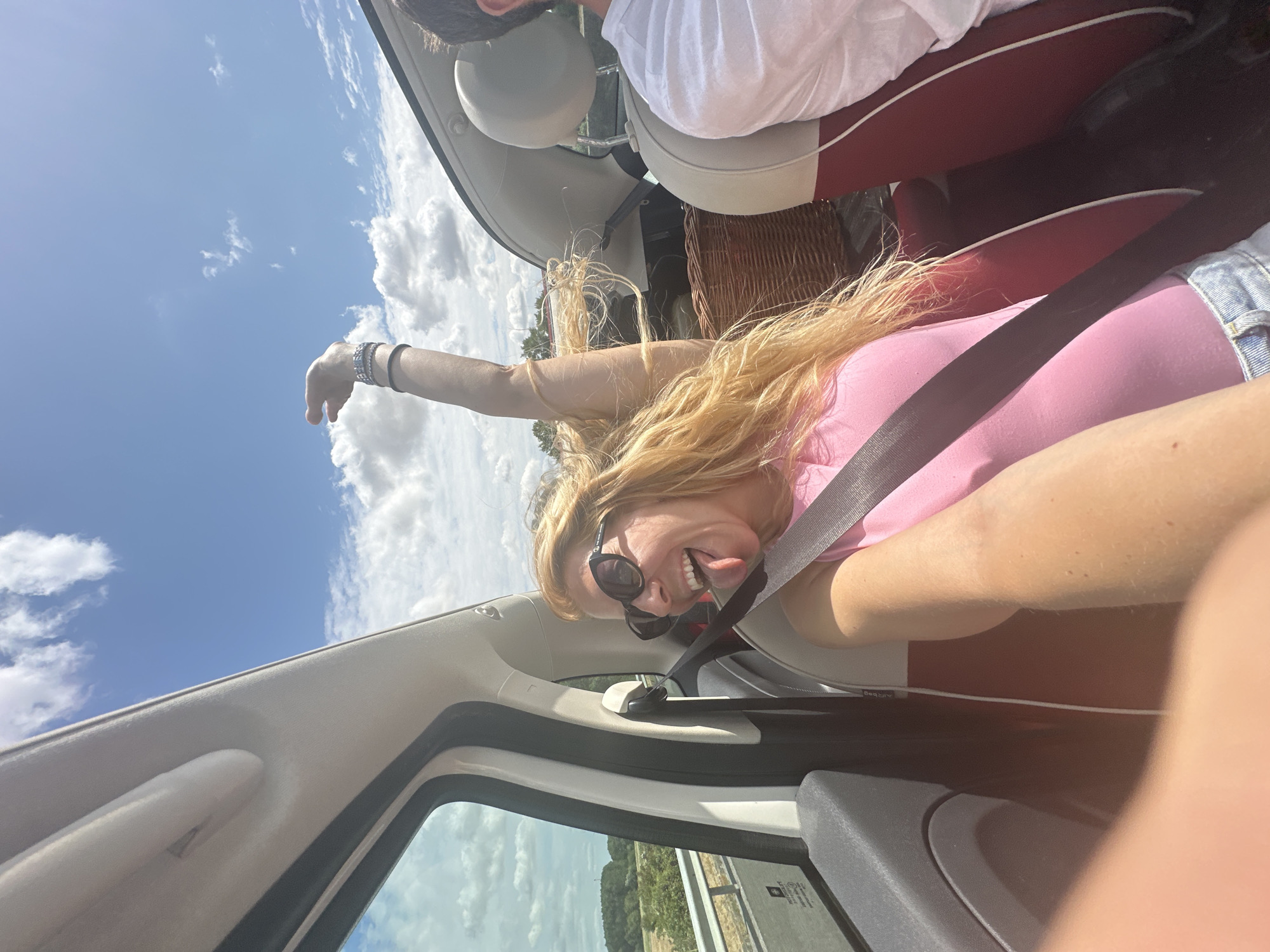 A blonde woman in a pink top and denim shorts sticks her tongue out at the camera while riding in a convertible car. She wears sunglasses and several bracelets on her arm. Her arm is raised in the air. In the background, a man in a white shirt is visible in the passenger seat. The car appears to be on a highway, with a landscape of trees and fields visible outside the window. The sky is blue with scattered clouds. The image is a selfie taken from the perspective of the driver or front passenger.