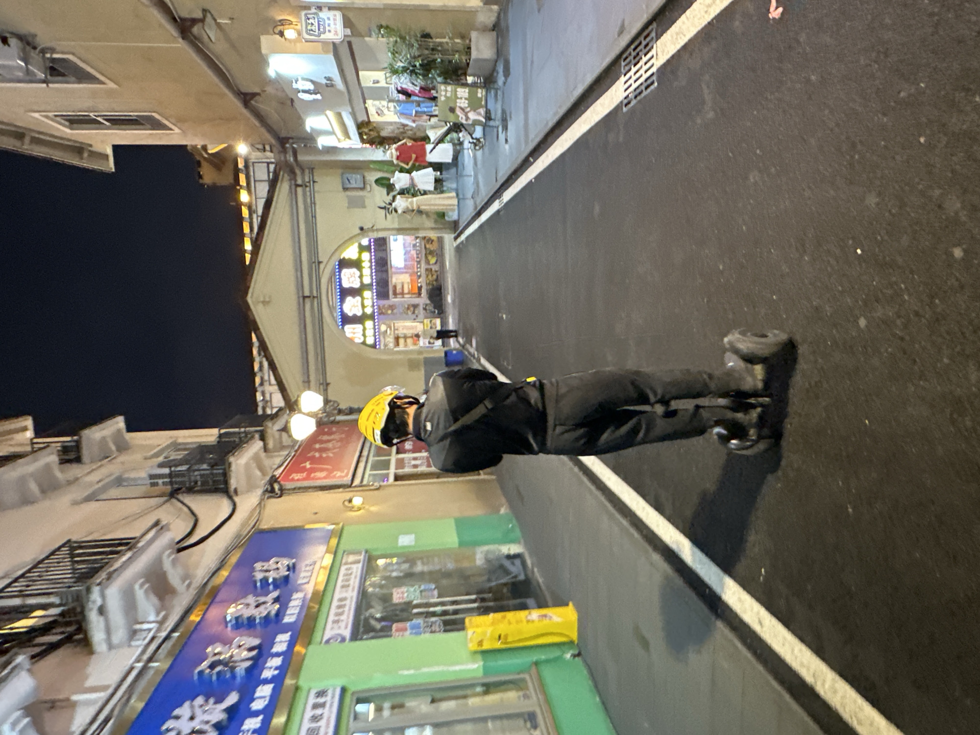 In the city of Chongqing High-tech Zone, China, an individual wearing a black jacket, black pants, and a yellow helmet is riding a self-balancing scooter down a paved street at night. The street is lined with buildings on both sides. On the left, a bright green storefront has a large blue sign with Chinese characters and a gold border. To the right of the green building, a red sign with white Chinese characters is visible. Further down the street, an arched entryway leads to an area with illuminated signs. Mannequins are displayed outside some of the shops. The street has white markings and a storm drain grate on the right side.