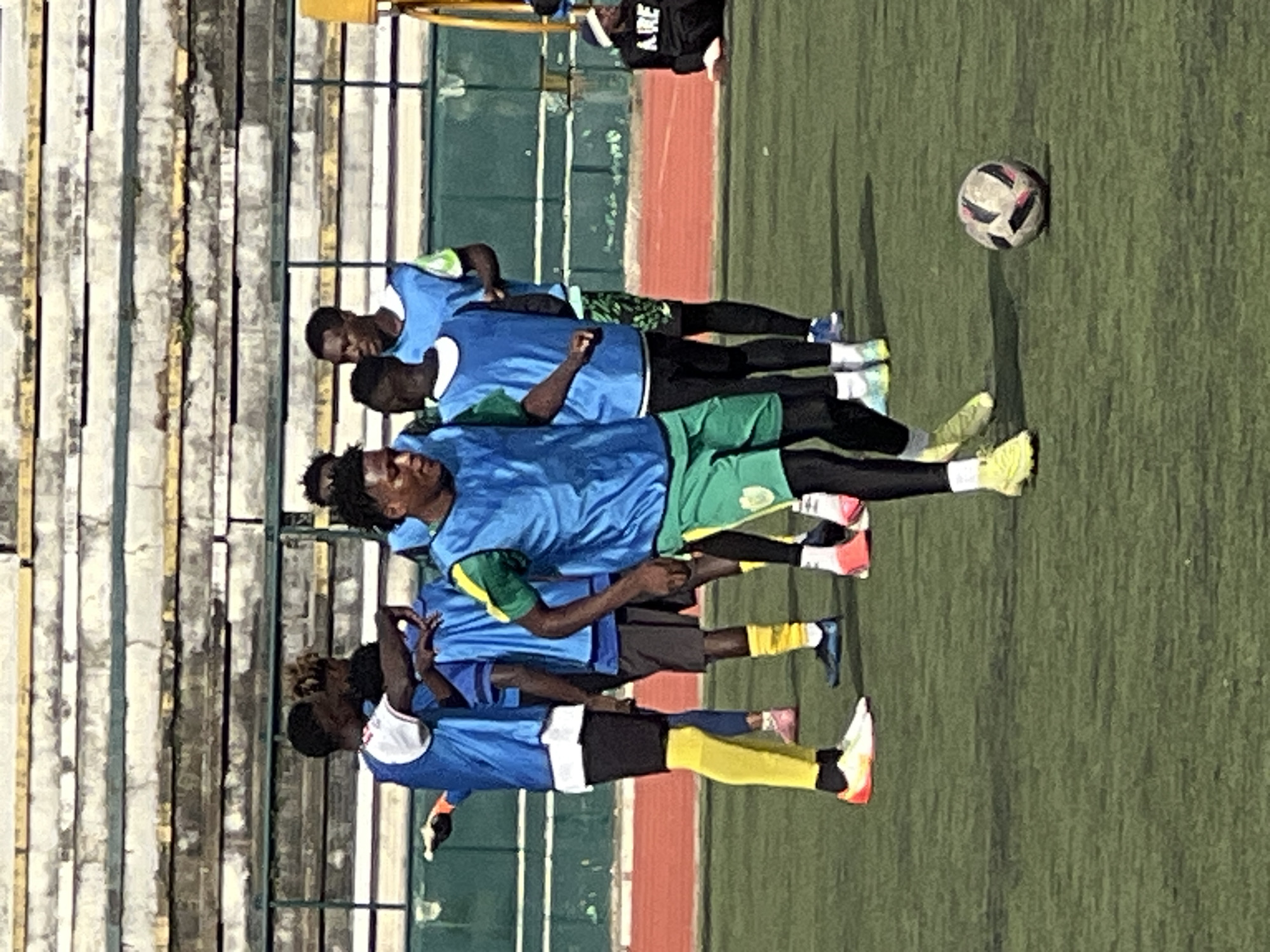 The image depicts a group of male soccer players on a green turf field, likely a practice session at the New Stadium in Nigeria. The players are wearing blue training bibs over their uniforms, with some also in green jerseys. One player in the foreground is actively dribbling a soccer ball. Behind the players, a set of bleachers with weathered concrete and yellow accents is visible, suggesting a well-used sports venue. The overall setting is outdoors under natural light.
