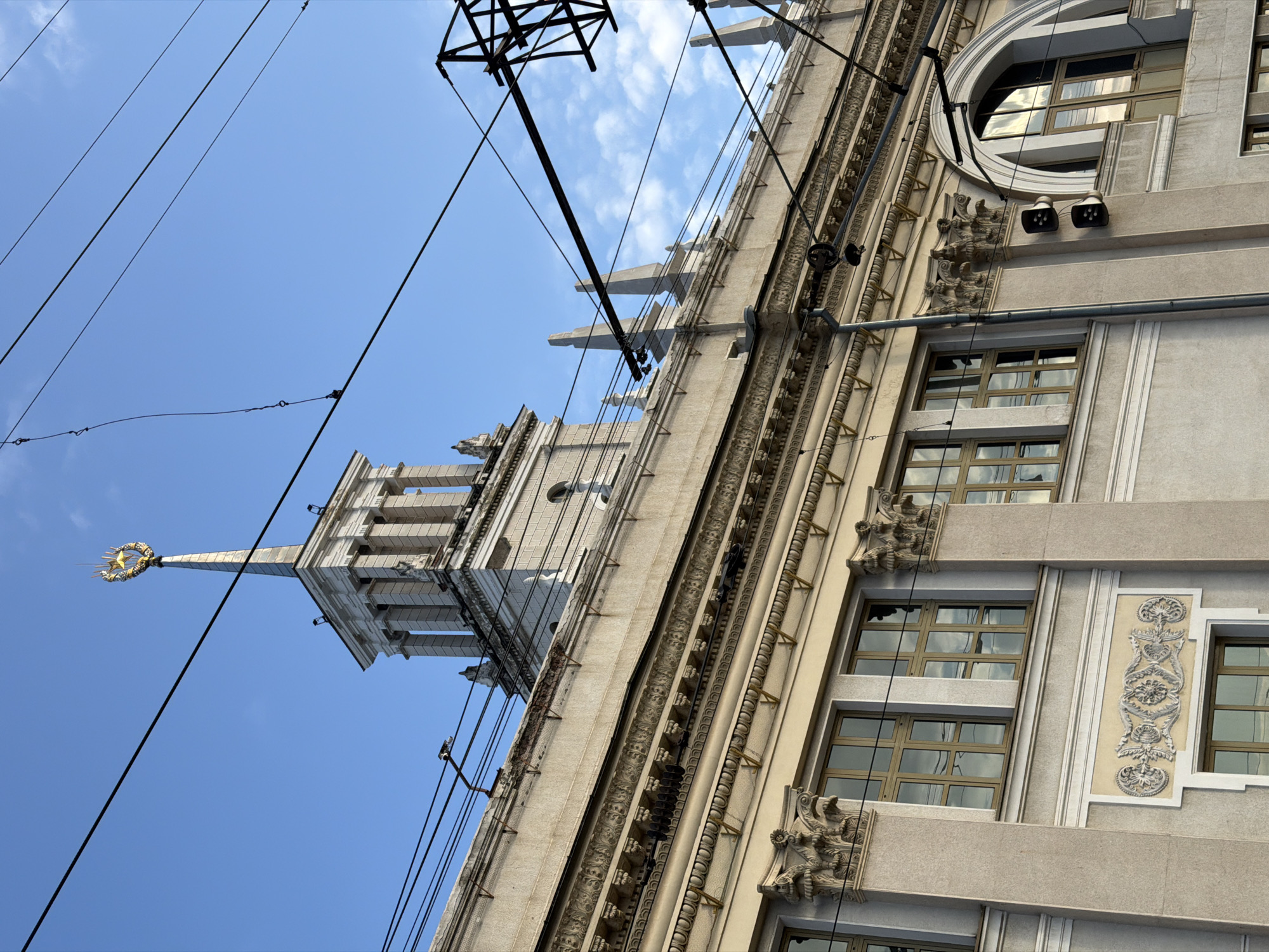 The image shows a low-angle view of a building in Krasnodar, Russia, likely the building at Krasnaya street 35. The building is light-colored, with ornate architectural details, including decorative cornices, window frames, and a tower topped with a spire. A golden star and wreath adorn the spire. Several windows are visible, reflecting the sky. Overhead power lines and a support structure are visible in the sky. The sky is a clear blue with some white clouds. The image is taken from a street-level perspective.