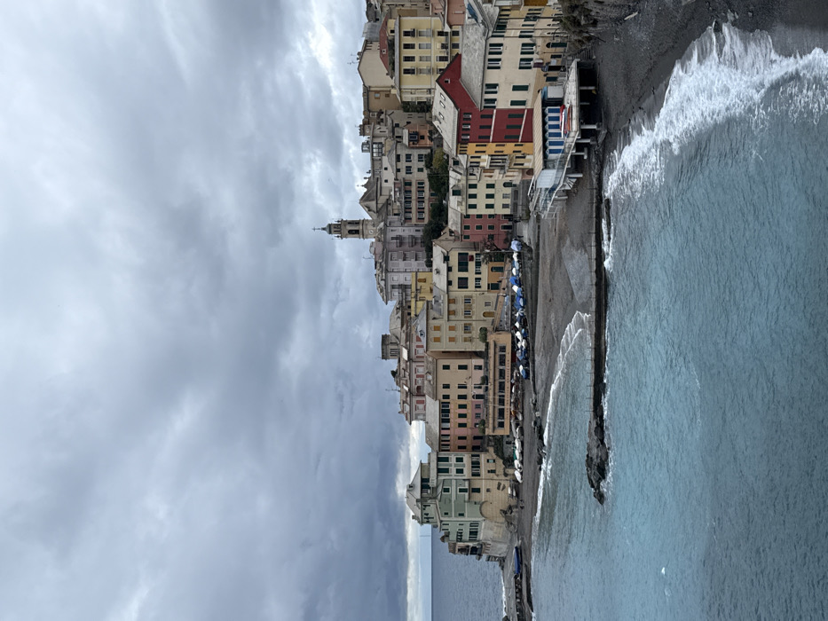 An aerial perspective captures the town of Bogliasco, Italy, under an overcast sky. The scene depicts a densely built coastal settlement with pastel-colored multi-story buildings, characteristic of Ligurian architecture, arrayed along a curved pebble beach and ascending a hillside. The Ligurian Sea, appearing turquoise near the shore and darker blue further out, displays small waves breaking on the dark grey beach. Two short, dark-rock breakwaters extend into the water.
Numerous small boats, many covered with blue or white tarpaulins, are stored on the beach in rows. A building with an orange awning, likely a restaurant, occupies a beachfront position on the left, with outdoor seating visible. To the right, a white building with blue shutters is elevated on pilings above the beach. A prominent bell tower (campanile) with a clock face and a spire rises at the highest point of the visible town, indicating a church. Scattered indistinct human figures are present on the beach and along walkways, appearing small due to the viewing distance. The overall environment is one of an active but quiet coastal town on a cloudy day.