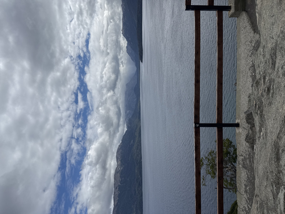 The image presents a panoramic outdoor landscape, captured on what appears to be a partly cloudy day. The scene is dominated by a large body of water, imposing mountains, and a dynamic sky, viewed from a vantage point.
In the foreground, on the right side of the frame, there is a sturdy, light grey, rough-textured stone or concrete wall. Attached to this wall is a dark, possibly metal, railing or fence structure. This railing consists of three visible vertical posts, evenly spaced, with two horizontal bars connecting them. A small tree with vibrant green foliage is partially visible peeking out from behind the railing near the bottom right corner, adding a touch of nature to the immediate foreground.
The midground is dominated by a vast body of water, likely a fjord or a large lake, which stretches horizontally across the image, filling the bottom and central portions. The water's surface is a muted blue-grey, calm but with subtle ripples visible, suggesting a gentle breeze or current. On the opposite side of this expansive water, a range of steep, dark grey to green mountains rises dramatically. These mountains are rugged and appear to be partially covered in dense vegetation, particularly at their bases and lower slopes. They recede into the distance, with some peaks appearing slightly hazy or obscured by clouds, contributing to the depth of the scene.
The background is filled with a dramatic sky. It is largely covered by a mixture of white and grey cumulus clouds, indicating a partly cloudy day. However, a distinct strip of clear blue sky cuts horizontally through the clouds, providing a contrasting band of vibrant color. The clouds vary in density and form, some appearing soft and fluffy, others more widespread and opaque.
There is no specific activity or event depicted, but the setup with the railing suggests this is a designated viewpoint, offering a breathtaking vista of the natural surroundings. The time of day appears to be daytime, with ample natural light illuminating the scene. No text is visible in the image. The overall impression is one of majestic natural beauty and serene grandeur.