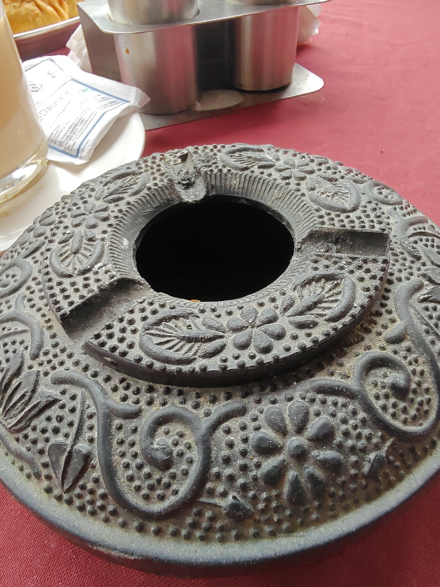 The image shows a close-up view of a decorative, dark-colored ashtray with floral and dotted patterns. The ashtray has a central opening and a notched edge, likely for resting cigarettes. It is placed on a red tablecloth. In the background, a glass containing a light-colored beverage, a white paper packet with blue text, and a stainless steel container are visible. The scene appears to be set in an outdoor dining area, possibly in Vashisht, India, given the cultural context suggested by the items and decor.
