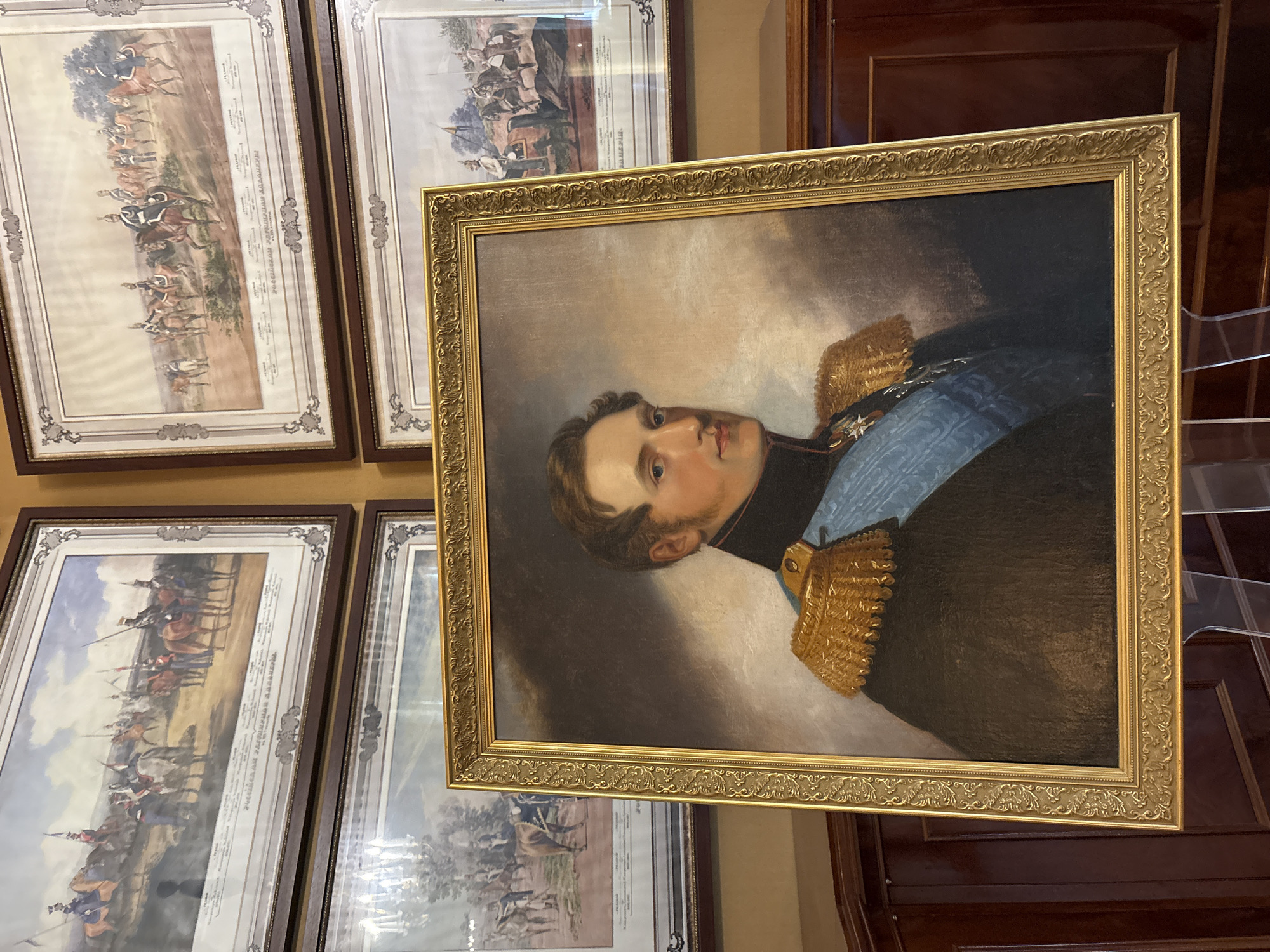 The image shows a framed portrait of a man wearing a military uniform with gold epaulets and a blue sash, set against a dark background. The portrait is a central focal point. Behind and to the sides of the portrait, other framed artworks are visible, depicting cavalry scenes. The overall setting appears to be an interior space with wooden cabinetry. This portrait is housed in the Gatchina Palace in Gatchina, Russia, specifically in the Emperor Alexander III Museum, part of the Gatchina State Museum-Reserve. The specific room is the Emperor's study.
