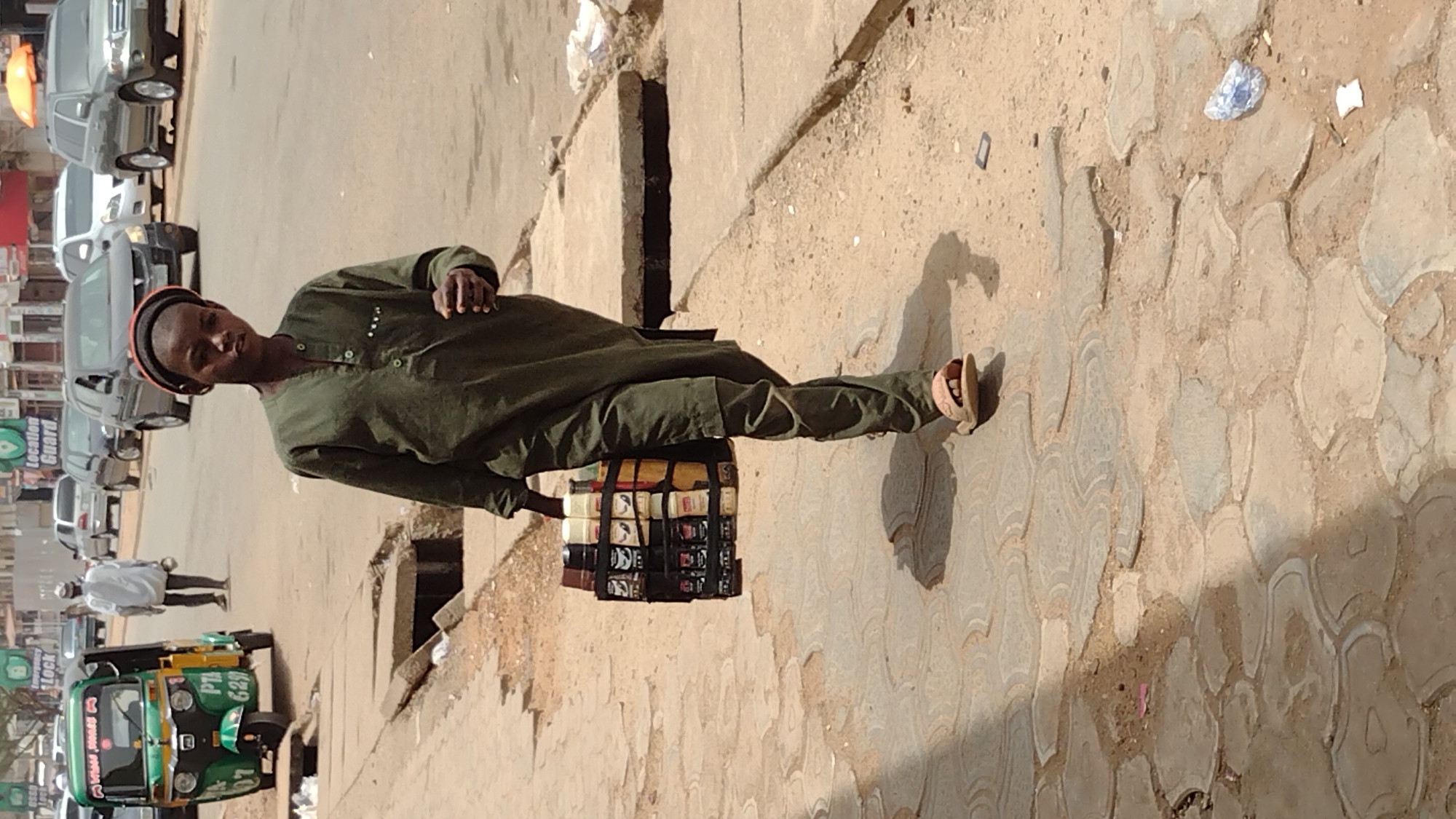A young man in Jos, Nigeria stands on a stone-paved sidewalk beside a city street. He is wearing a dark green buttoned tunic and trousers with brown sandals. He is carrying a stack of what appears to be books or notebooks secured together with straps. The street next to the sidewalk contains a green and yellow auto rickshaw in the foreground, and multiple cars behind it. There is some litter strewn on the sidewalk.