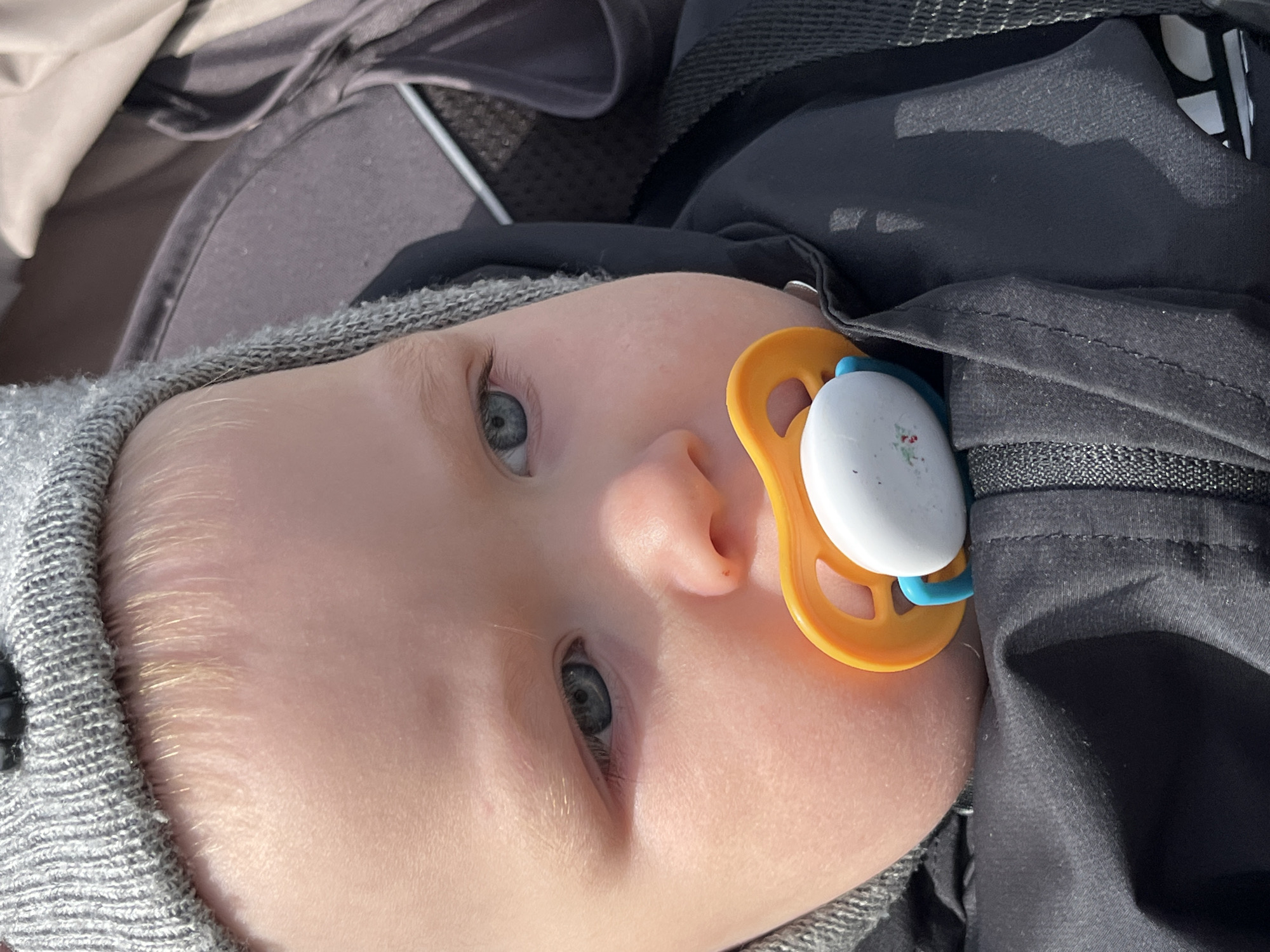 This close-up, rotated image features a baby lying in what appears to be a stroller or carrier, captured during daylight in Chaikovsky, Russia. The baby has fair skin, light blue-grey eyes, and fine blonde hair peeking from under a grey knitted beanie. A neutral, calm expression is on the baby's face, with eyes open and looking slightly towards the upper right.
The baby is dressed in a warm grey knitted hat and a dark grey or black hooded jacket, indicating cooler weather conditions. An orange and white pacifier is in the baby's mouth, with the white part adorned by faint red and green speckles, connected by a light blue loop. Bright sunlight illuminates the left side of the baby's face, creating soft shadows on the right.
The background shows elements of a dark grey fabric stroller or carrier, including a metal frame. The attire suggests an outdoor setting during a cooler season like autumn, spring, or a mild winter day. No discernible text is visible on any of the objects or clothing.