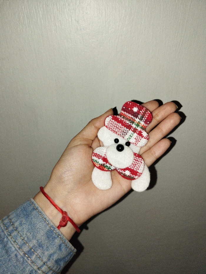 A hand holds a white plush toy shaped like a dog or bear with a red and green plaid pattern on its hat and body. The toy has two small, black bead eyes. The hand belongs to someone wearing a denim jacket with a red string bracelet. The background is a plain light gray wall, casting a slight shadow of the toy onto the palm.