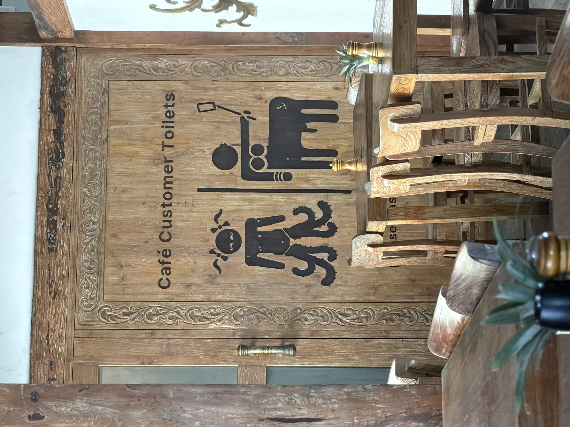 The image shows a carved wooden door in Unknown, Indonesia. The door is decorated with carvings around the perimeter and features a sign indicating "Café Customer Toilets." The sign uses stylized black silhouettes to represent male and female figures, differing in their body shapes and attire. The male figure is depicted with what appears to be a wheelchair symbol or assisted device, with the female figure represented with tentacles and facial hair. The door has a simple metal handle and appears to be part of an establishment like a cafe or restaurant. There are several chairs beside the door. A small plant on the chair beside the door.