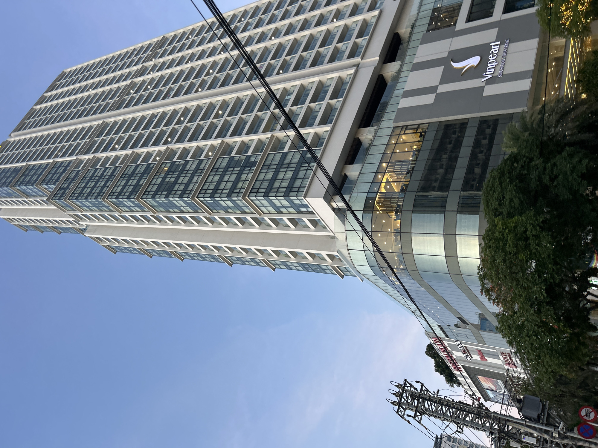This is a ground-level, eye-level, medium shot photograph of a modern skyscraper in Nha Trang, Vietnam. The skyscraper, identified by the "Vinpearl Beachfront Nha Trang" logo on its lower section, dominates the frame, extending upwards into a clear blue sky. The building features a white facade with numerous balconies and windows, giving it a sleek, contemporary appearance. The lower portion of the building is encased in a curved glass structure, revealing interior lighting and people. Adjacent to the skyscraper, a utilitarian electrical pole with numerous wires is visible on the left side of the image, along with some street signage. Green trees partially obscure the base of the buildings. The overall scene suggests an urban environment.