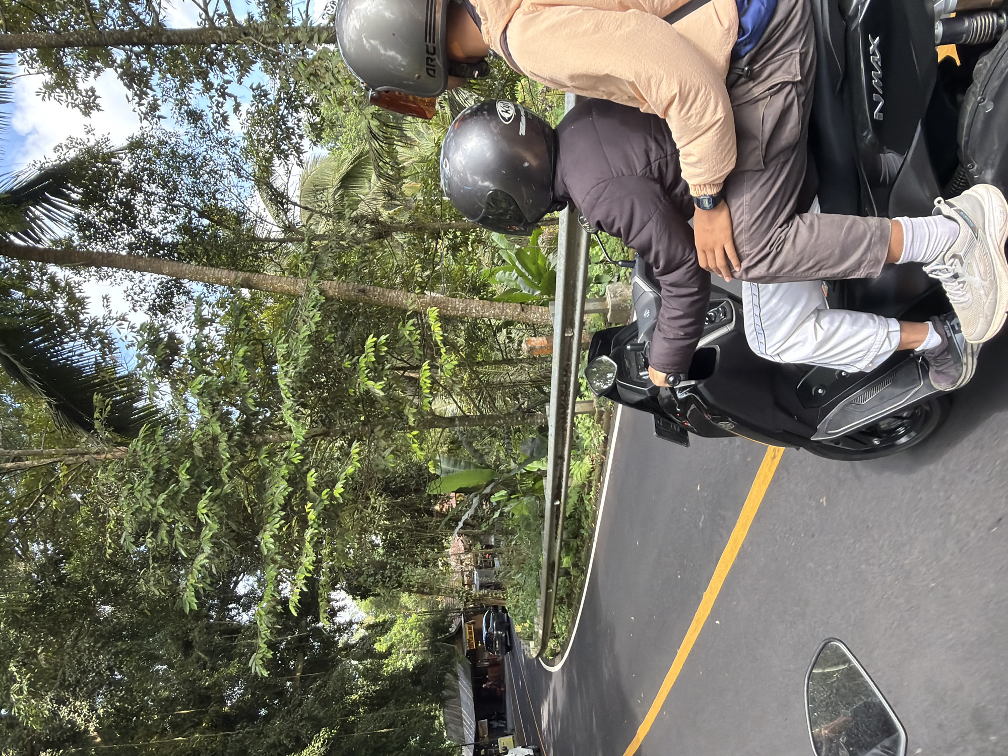 Two individuals are riding a black Yamaha NMAX scooter on a paved road in Tampaksiring, Indonesia. The scooter has a yellow line marking on the asphalt to its left. The riders, both wearing helmets, are positioned one behind the other. The person in front is wearing a dark jacket and white pants with a blue stripe, and the person behind is wearing a light peach-colored jacket and brown pants. The environment is lush and green with tall trees lining the road, suggesting a rural or tropical setting. Buildings with traditional architecture are visible in the background, situated on the side of the road. The sky is partly cloudy.