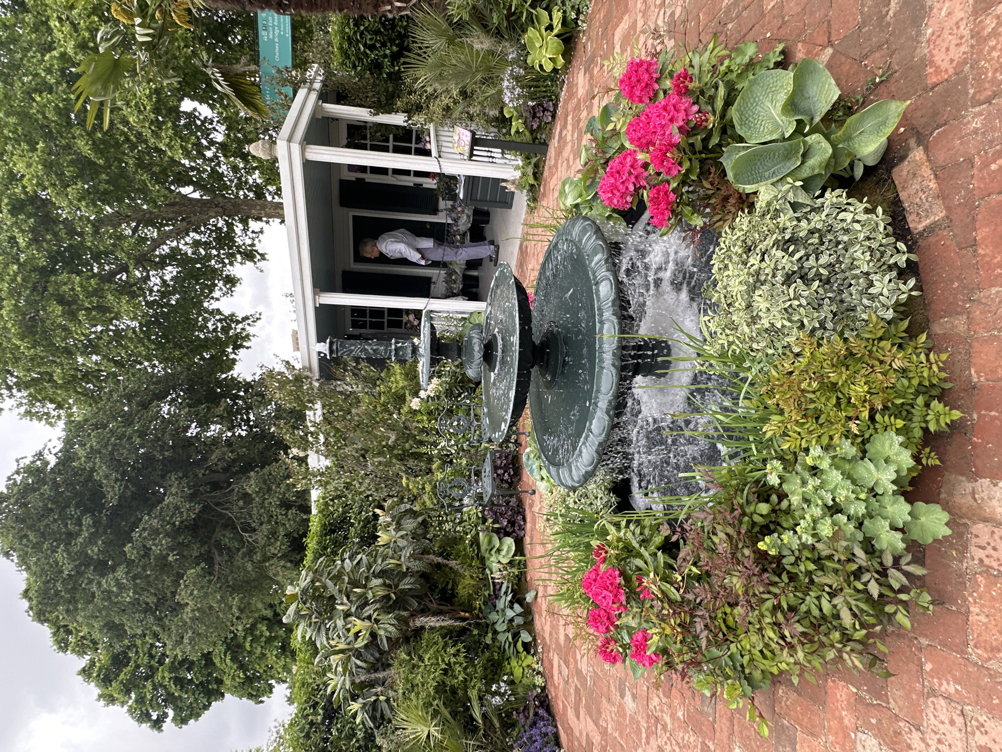 A fountain surrounded by plants and flowers sits in front of a building on a brick path. A person walks into the building, which is likely in London, United Kingdom. The image captures a garden setting with a two-tiered fountain as the focal point. The fountain is positioned amidst a variety of plants and flowers. A brick path leads to a building with columns. A person is entering the building. Trees are visible behind the building. The sky is overcast. A sign indicating "Chelsea Bridge Road" can be seen, suggesting the location is likely in London, United Kingdom.