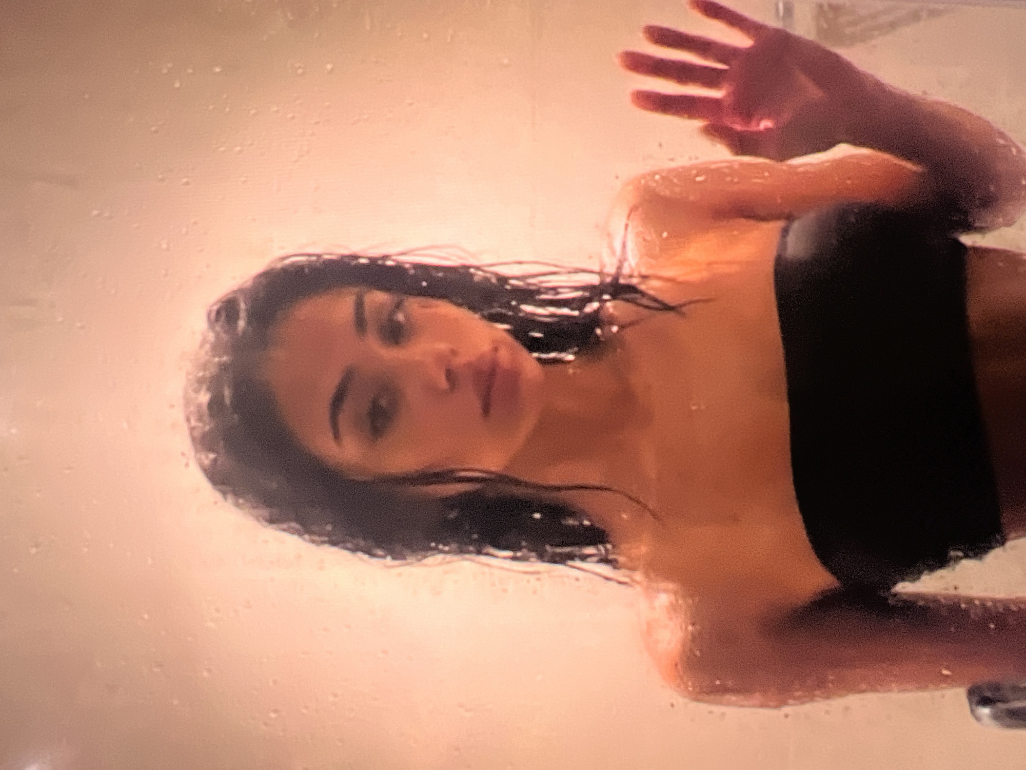 A young woman with wet, dark hair and a black strapless top stands behind a surface with water droplets. She is looking at the camera with her right hand raised and pressed against the surface. The background is a blurred pinkish-orange hue.