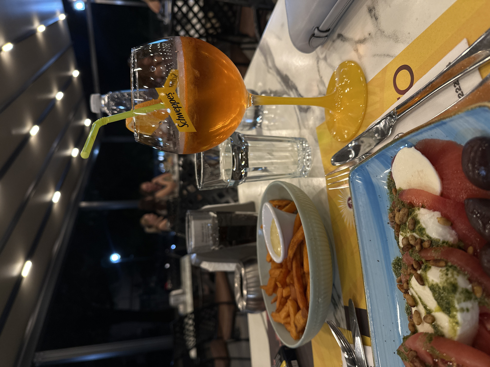 The image depicts an outdoor dining scene at night, likely at a restaurant in Petrich, Bulgaria. In the foreground, a table is set with food and drinks. A bright orange beverage, possibly an Aperol Spritz, is served in a tall stemmed glass with a green straw and an orange slice. Beside it, a glass of water and a portion of sweet potato fries with a dipping sauce are visible. A blue plate holds a Caprese salad with tomatoes, mozzarella, basil pesto, pine nuts, and olives. Utensils, including a fork and knife, rest on the table, along with a grey smartphone case. In the background, indistinct figures of people are seated at other tables, suggesting a lively atmosphere. The setting is under a covered patio with overhead lighting.