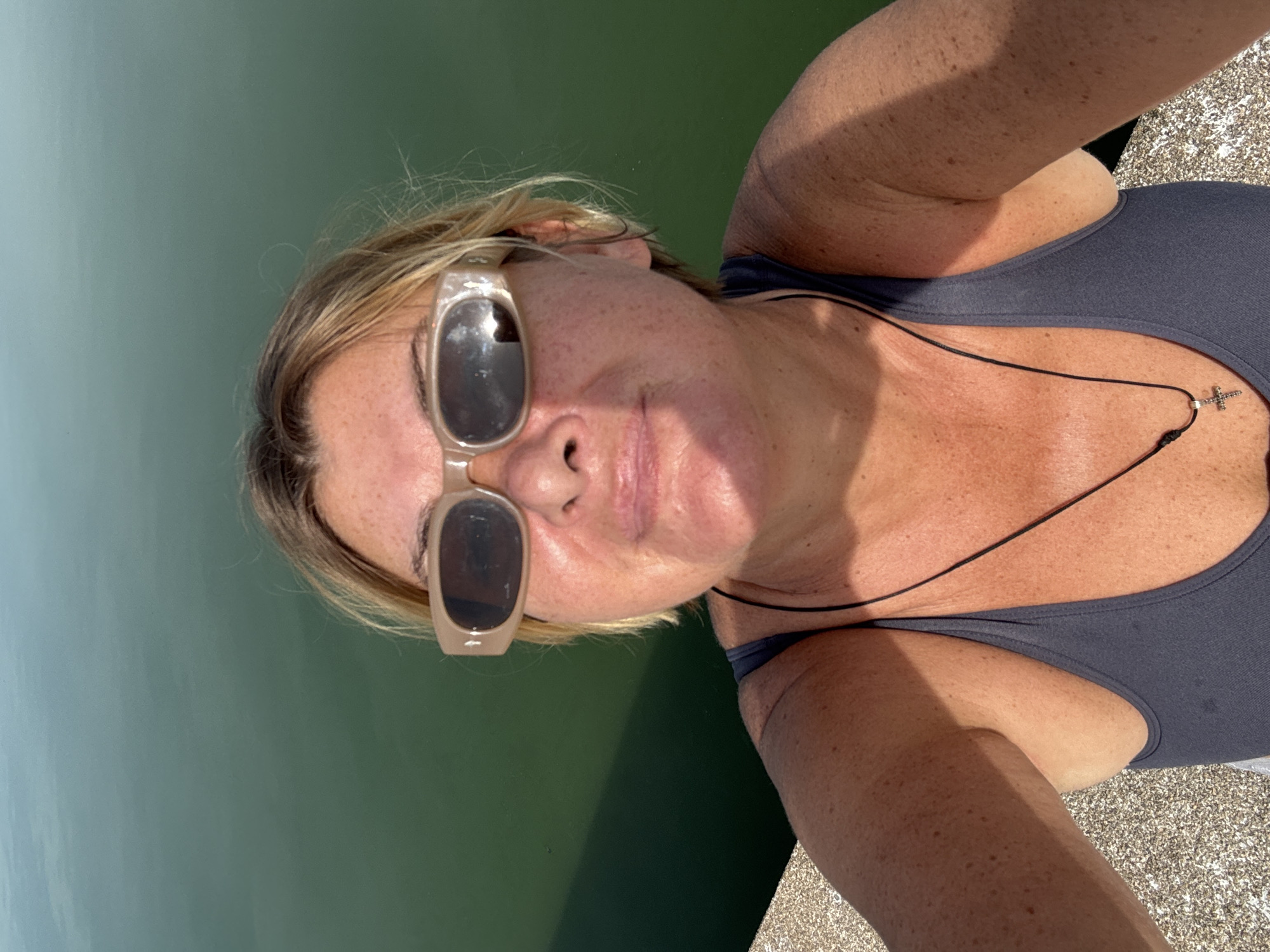 A woman with blonde hair and sunglasses is taking a selfie outdoors. She is wearing a dark tank top and a cross necklace. The background is a body of green water, possibly a lake or ocean, with a concrete ledge in the foreground. The lighting suggests it is daytime and sunny.