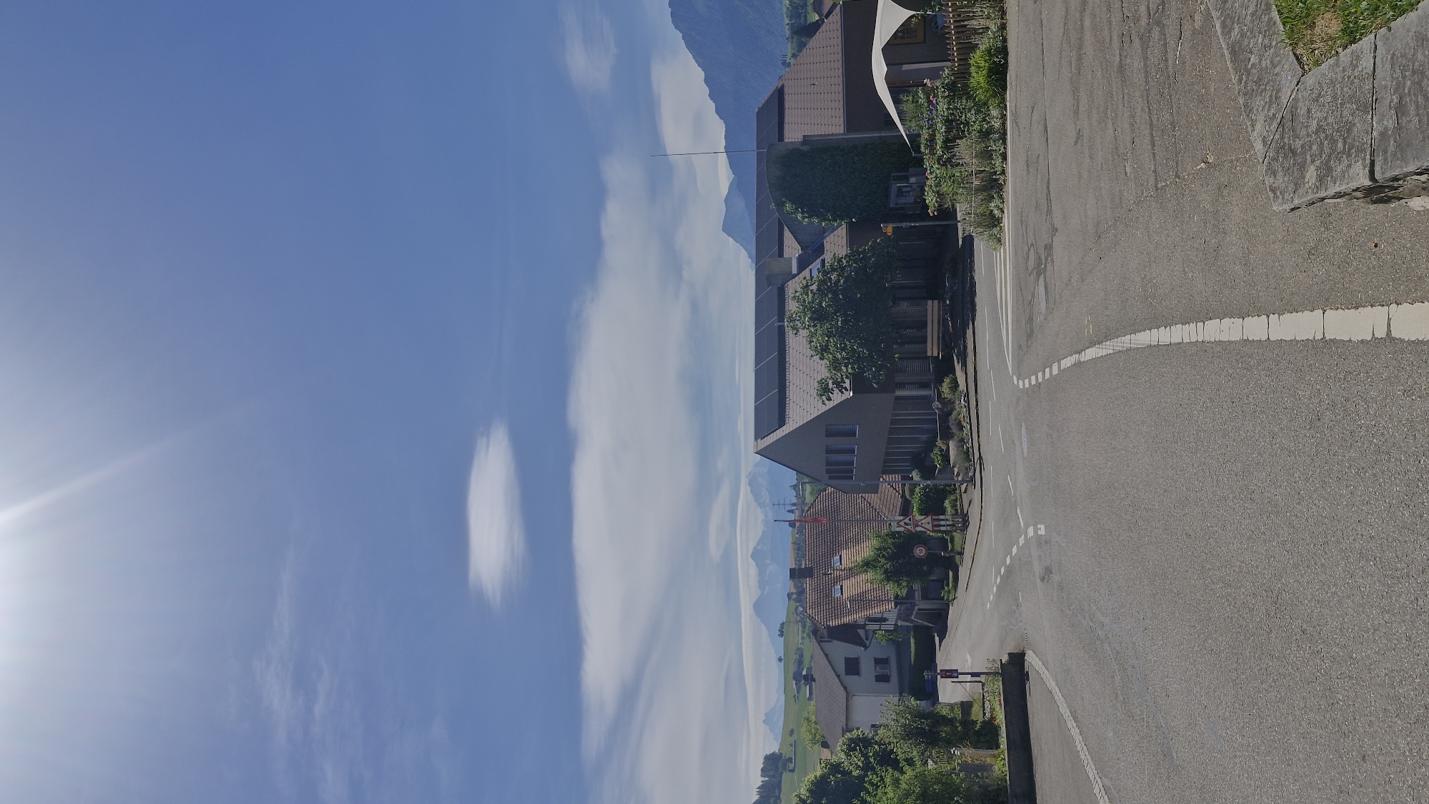 The image depicts a sunlit street scene in Wattenwil, Switzerland. The road, paved with asphalt, curves through a residential area lined with houses. White dashed lines mark the lanes of the road, indicating traffic direction. Some houses feature modern architecture with large windows and dark roofing, while others have more traditional tile roofs. Lush green vegetation, including trees and bushes, surrounds the buildings. In the distance, majestic mountains are visible, partially obscured by wispy white clouds. The sky is a vibrant blue, with the sun's rays creating a bright glare on the left side of the image. The time of day appears to be midday or early afternoon, given the bright sunlight and shadows. There are no people or specific activities immediately apparent in the image, suggesting a quiet, everyday scene. The overall impression is one of a peaceful and scenic residential area within a mountainous landscape. A road sign with the number "50" is visible, likely indicating a speed limit.