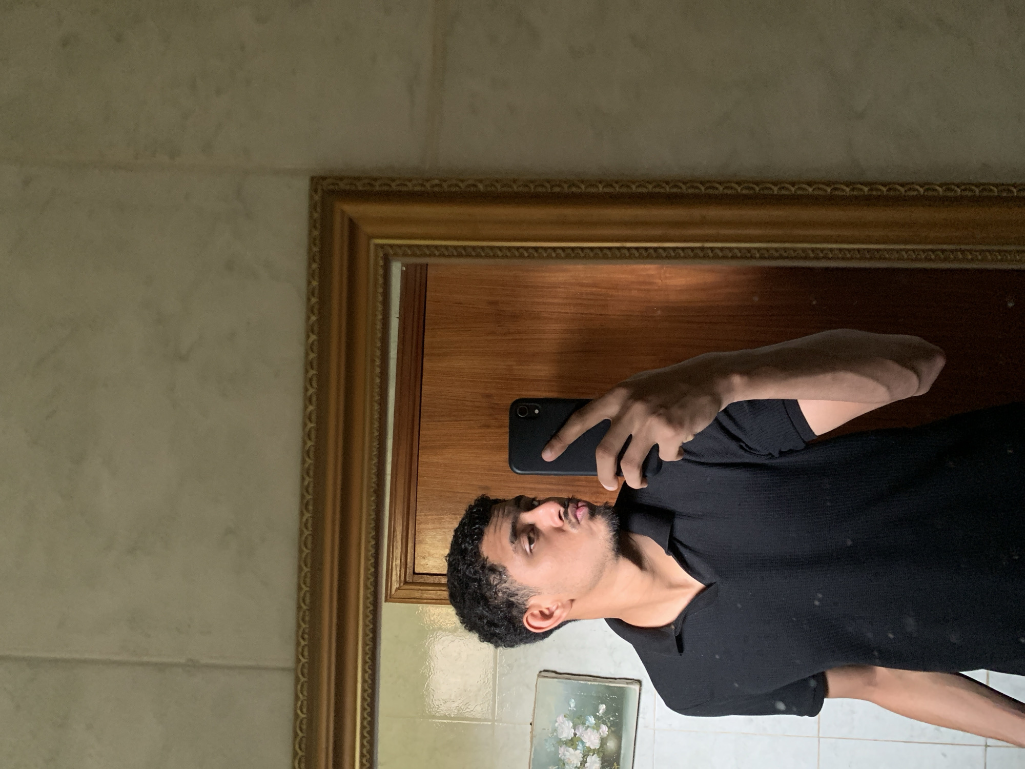 An indoor mirror selfie captures a man taking a photograph of himself with a black smartphone. The image is oriented vertically, showing the man from the chest up, reflected in a mirror with a decorative golden frame. He is looking slightly upwards and to his right, towards the camera of his phone, which he holds in his right hand. He has dark, curly hair, a beard, and is wearing a black collared shirt.
The background reflected in the mirror appears to be a light-colored wooden door or panel. Below the man's reflection, a small framed picture or painting with light blue tones and white flowers is visible on what looks like a tiled wall. The wall surrounding the mirror is covered in light-colored, possibly beige or off-white, textured tiles or wallpaper. The overall lighting is soft, suggesting an indoor environment without direct harsh sunlight. There is no visible text in the image. This scene is set in Santa Ana, Venezuela.