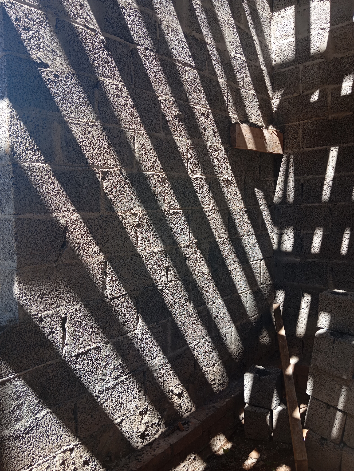 The image depicts a rough, unfinished concrete block wall with strong diagonal shadows cast across it, suggesting the presence of a nearby slatted structure or window. The wall appears to be part of a construction site or an abandoned building, with visible mortar lines and the textured surface of the blocks. There are a few scattered pieces of wood and stacks of concrete blocks visible in the lower right portion of the frame, further indicating a construction or renovation context. Sunlight is clearly a dominant element, creating dramatic contrasts between light and shadow. The scene is indoors or in a partially enclosed space, as evidenced by the walls and the limited view of the exterior. There are no people visible in the image, and no specific actions are being performed. The environment is dusty and appears unmaintained. The location is identified as being in Orenburg, Russia.