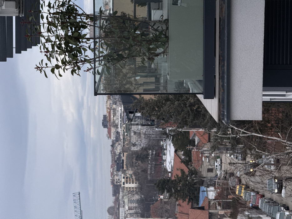 A cityscape with a cloudy sky is viewed from above. Balconies with glass panels are visible, one with a potted plant. The city consists of a mix of low-rise buildings with red roofs and taller structures, with some greenery interspersed. A construction crane appears on the left. A mix of overcast weather and residential or commercial structures dominates the scene.