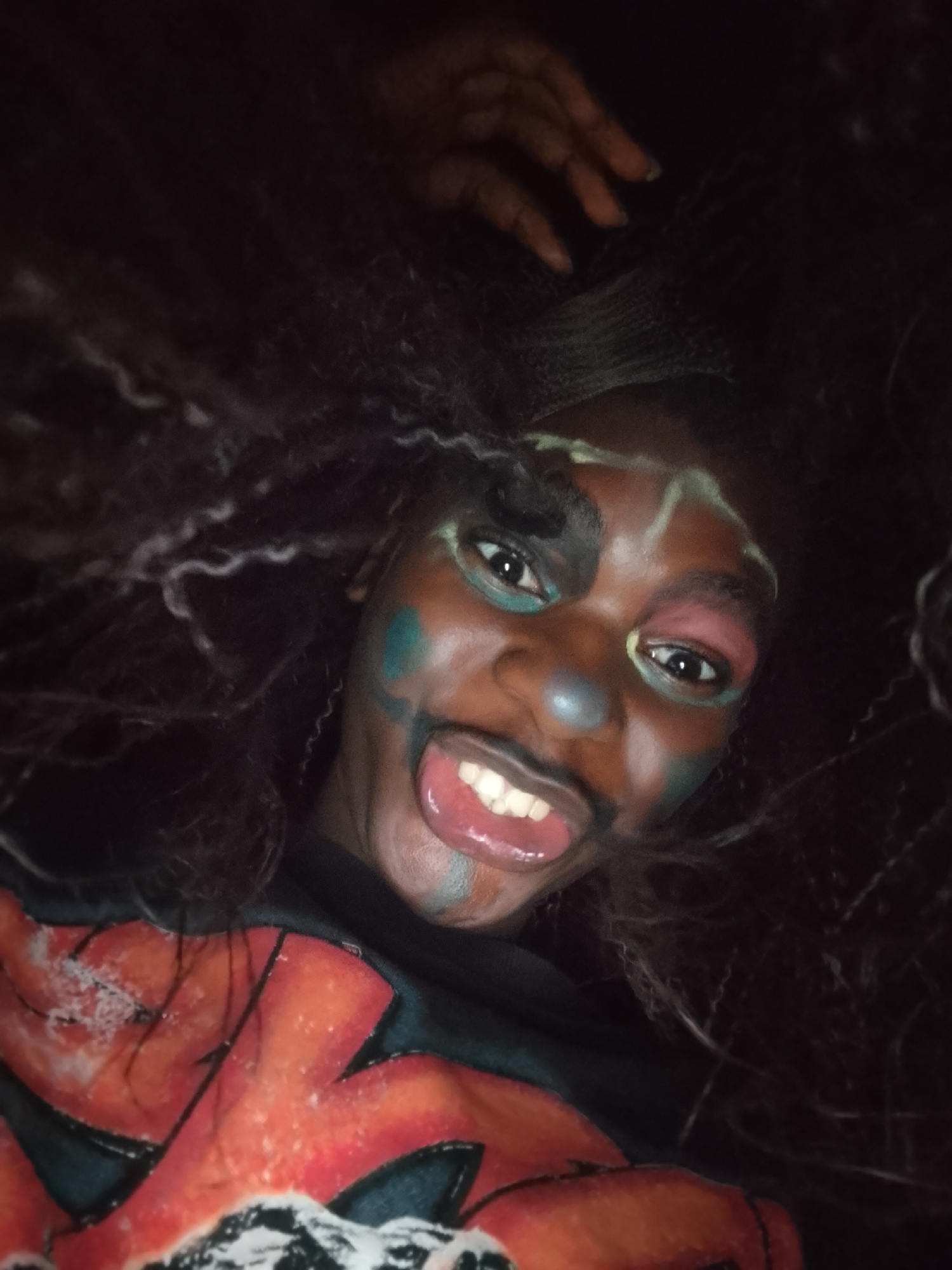 A close-up, dimly lit portrait captures a person with dark skin and elaborate, multi-colored face paint, looking directly at the camera with a playful expression. The face is adorned with bold black makeup around the left eye, extending towards the temple, alongside vibrant green streaks on the left cheek and chin. A distinct light blue dot sits on the nose, while pink makeup colors the area around the right eye, complemented by lighter highlights on the forehead and across the nose bridge.
The person displays a wide, open-mouthed smile, revealing white teeth and a portion of their tongue, conveying a sense of joy and high energy. Their dark, voluminous hair, styled in dense curls or braids, frames the face and falls around them. They are wearing a dark shirt featuring a prominent graphic design in bright orange and white. Part of their right hand is visible in the upper right corner of the frame, partially obscured by their hair.
The low-light setting casts a soft glow on the face, creating a dramatic contrast against the dark background and suggesting an evening or indoor event. There is no discernible text visible in the image. The overall impression is one of expressive celebration or performance, possibly at a festival or costume party.