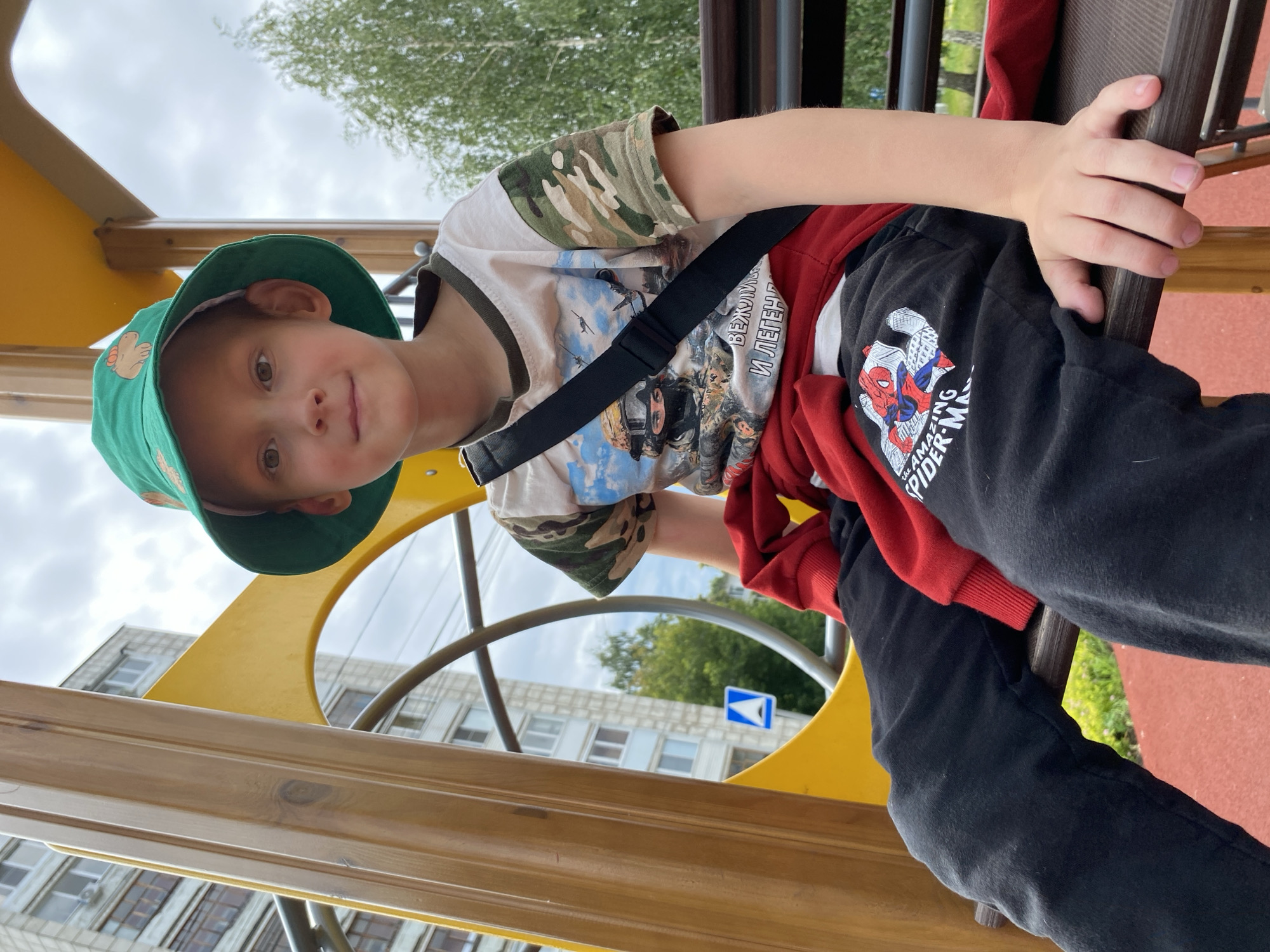 The media file shows a young boy in a green bucket hat and a camouflage-sleeved t-shirt sitting on a playground structure. He is wearing black pants with a graphic of Spider-Man on the left leg. A red hoodie is draped over his shoulders. His right hand is resting on a wooden railing. The boy is looking towards the camera with a slight smile.
The setting appears to be an outdoor playground with yellow and brown elements of the play equipment visible. In the background, there are trees, a cloudy sky, and a multi-story building that suggests an urban or suburban environment. A blue triangular sign with a white arrow is also visible in the background.
The weather appears to be overcast, indicated by the cloudy sky. The time of day is not explicitly stated, but the lighting suggests daytime. The overall mood is casual and relaxed. There is visible text on the boy's pants which reads "THE AMAZING SPIDER-MAN". The image's orientation is rotated, with the boy appearing to be upside down relative to a standard upright view. The location context is Chaikovsky, Russia.