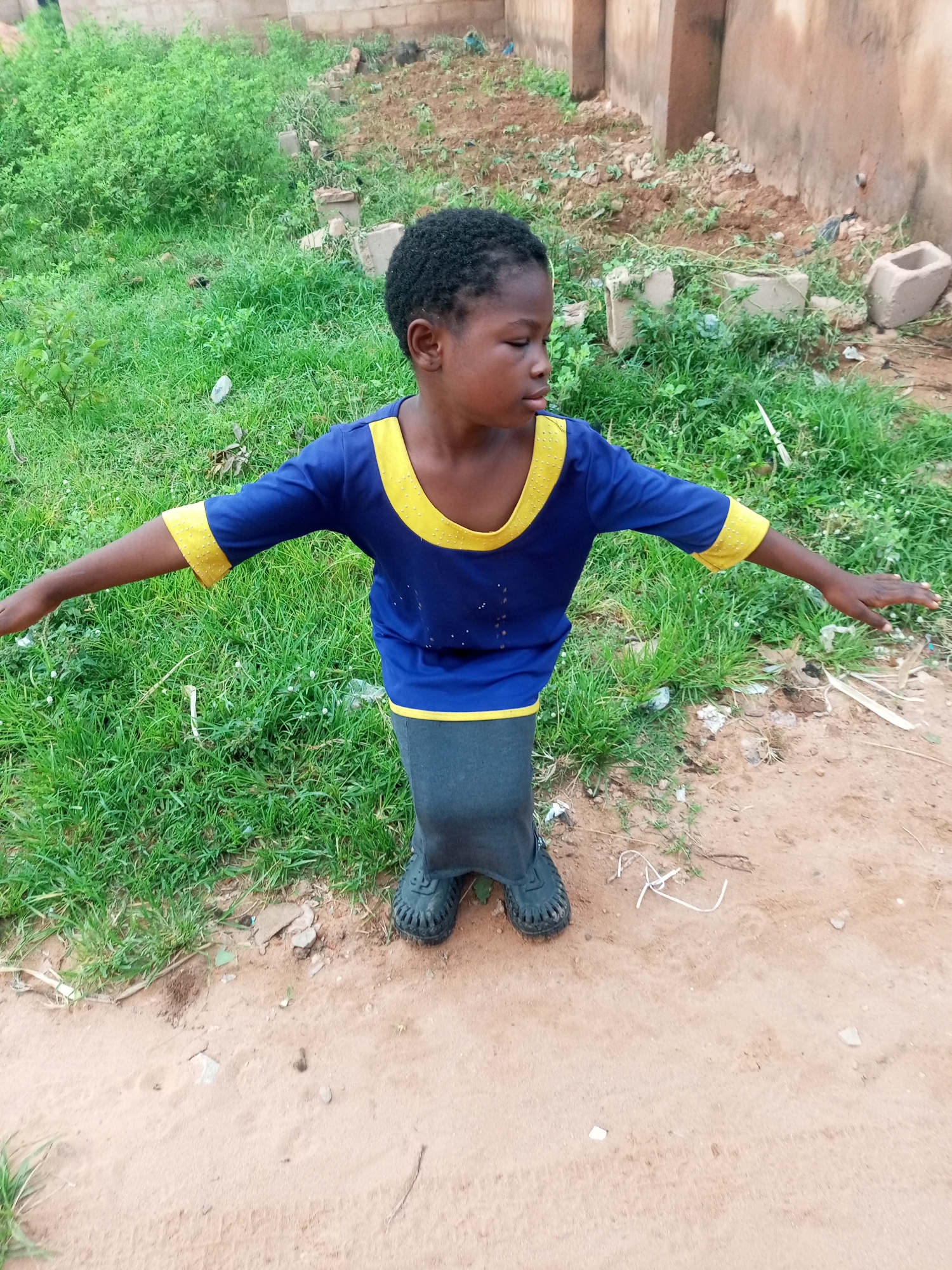 In Tempure, Nigeria, a young Black child stands outdoors, arms outstretched. The child wears a blue t-shirt with yellow trim and dark grey pants. The ground is a mix of sand and sparse green grass. In the background, a weathered brick wall and some construction debris are visible.