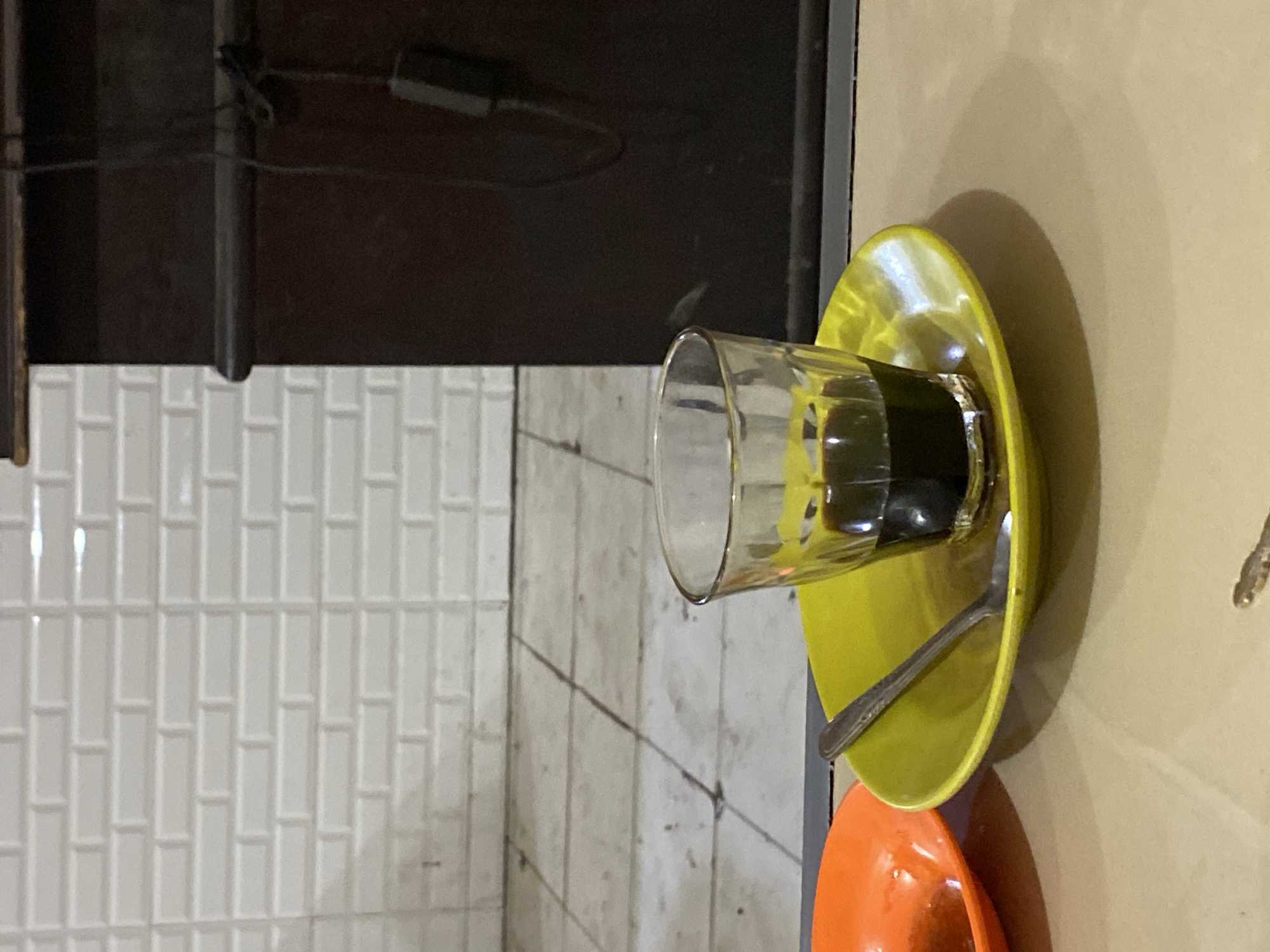 The image shows a glass of dark liquid, likely coffee, sitting on a yellow saucer. A silver spoon rests on the saucer. To the left of the yellow saucer, a portion of an orange saucer is visible. The background features white subway tiles and a dark wooden cabinet with a black cord plugged into it. The scene is set on a light brown table. The image appears to have been taken indoors, possibly in a cafe or home, and given the context of Lhokseumawe, Indonesia, it is likely a local establishment.