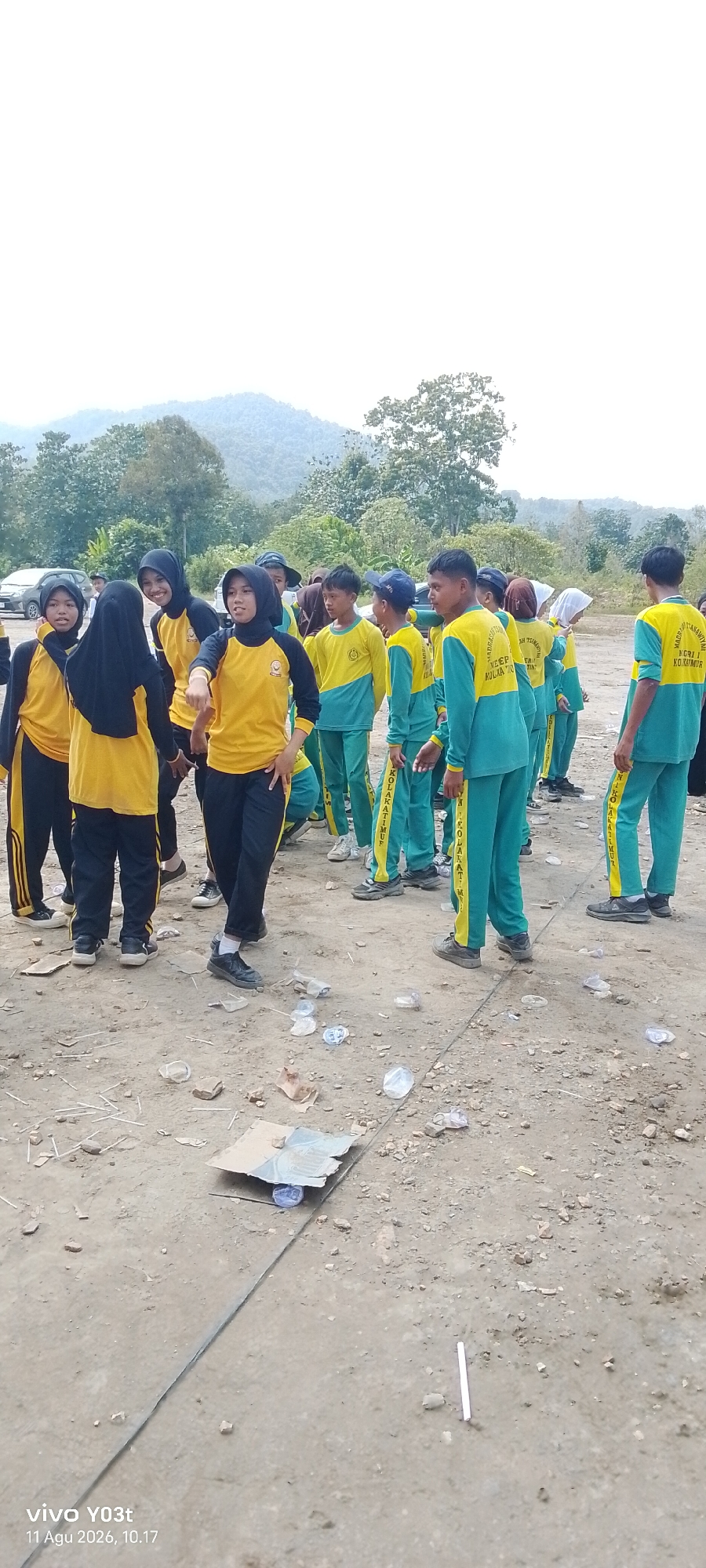 A group of students in yellow and green uniforms are gathered outdoors on a dirt ground, with a backdrop of hills and trees. The students appear to be in a school setting, possibly for an outdoor activity or event. Some students are standing in small groups, engaged in conversation, while others are looking in different directions. Scattered on the ground are plastic bottles, paper scraps, and small stones, indicating a somewhat unkempt environment. The presence of cars in the background suggests this location is accessible by road. The image is dated "11 Agu 2026, 10.17", indicating a time of morning. This scene likely depicts a school in Tirawuta, Indonesia, given the reference to the city in the prompt.