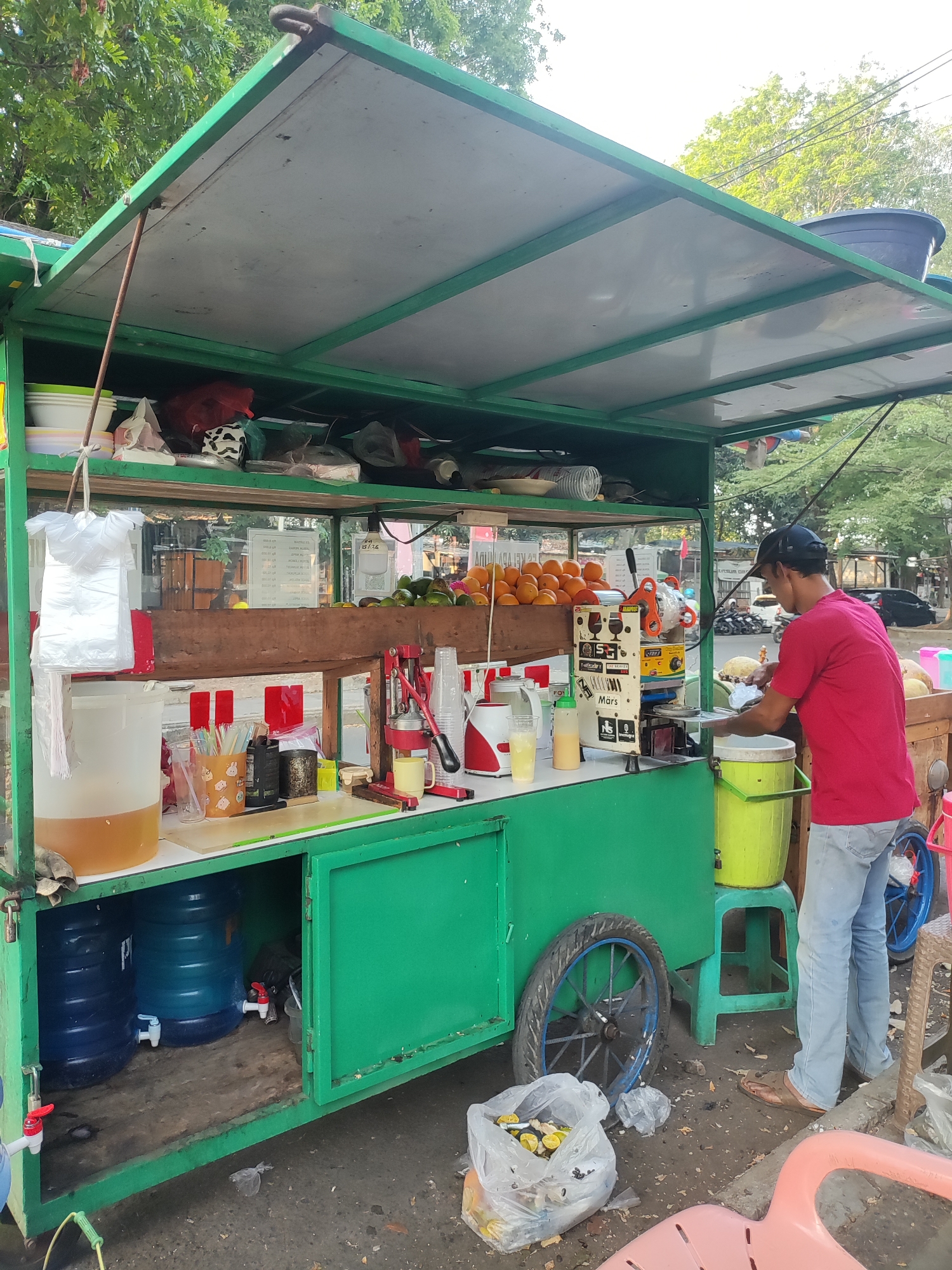 This image captures a street-side juice cart in Cirebon, Indonesia. A man in a red shirt and blue jeans is standing at the cart, preparing a drink. The cart is green and has a roof extending over the workspace. On the cart's shelves are various items including fruit (oranges and avocados are visible), plastic bags, cups, a juice press, and large water dispensers. Two large blue water barrels are stored beneath the counter. The ground around the cart is paved and appears somewhat dirty, with scattered debris and plastic bags. In the background, trees and other street elements suggest an urban environment, possibly a roadside stall.