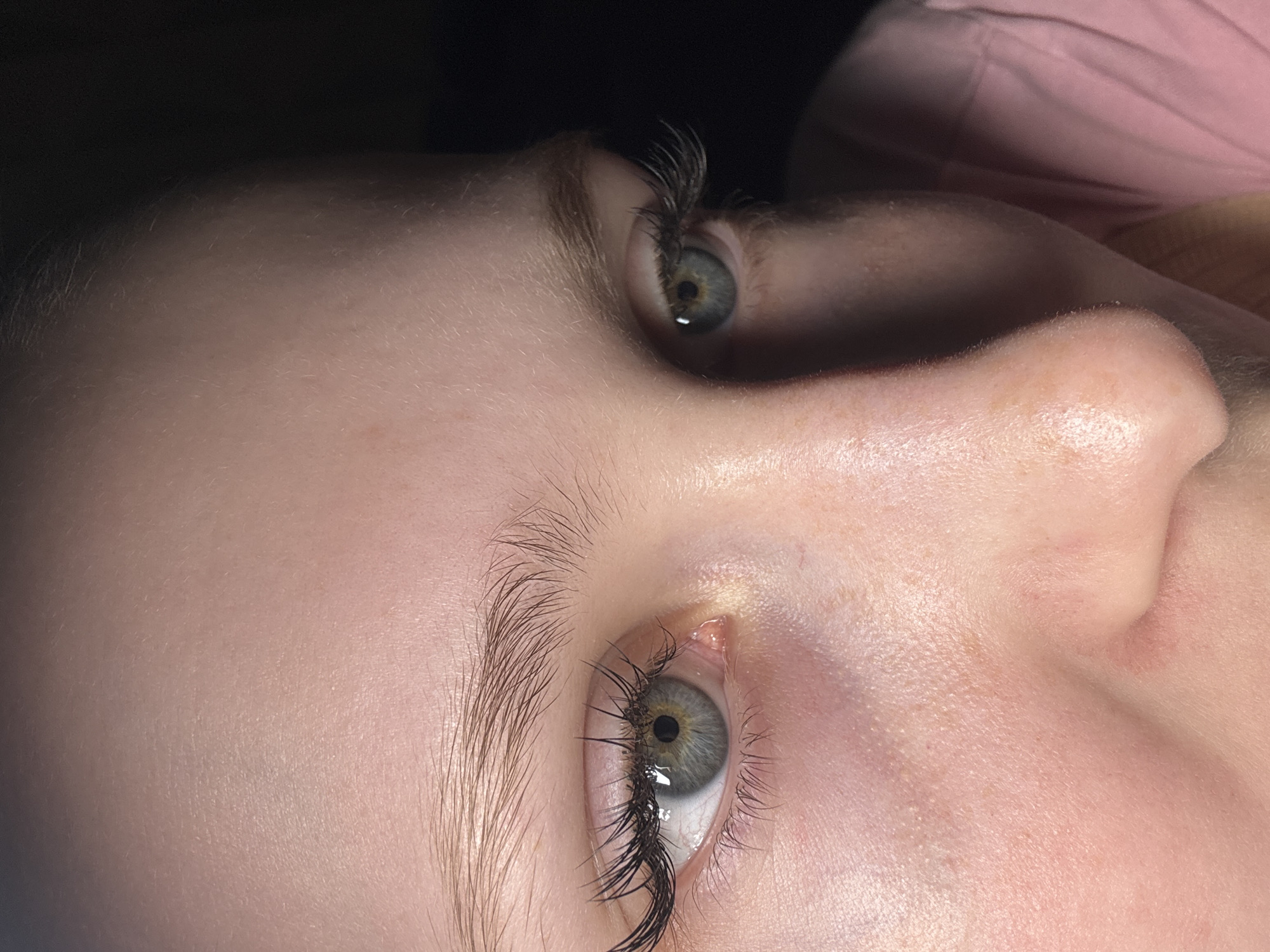 A close-up, angled shot focuses on a person's face, predominantly their eyes, nose, and forehead. The skin is pale, showing some freckles. The eyes are light-colored with dark lashes, which appear to be enhanced. The environment is dimly lit. The location and details are not discernible from the image to establish a connection to the city of Chaikovsky, Russia.