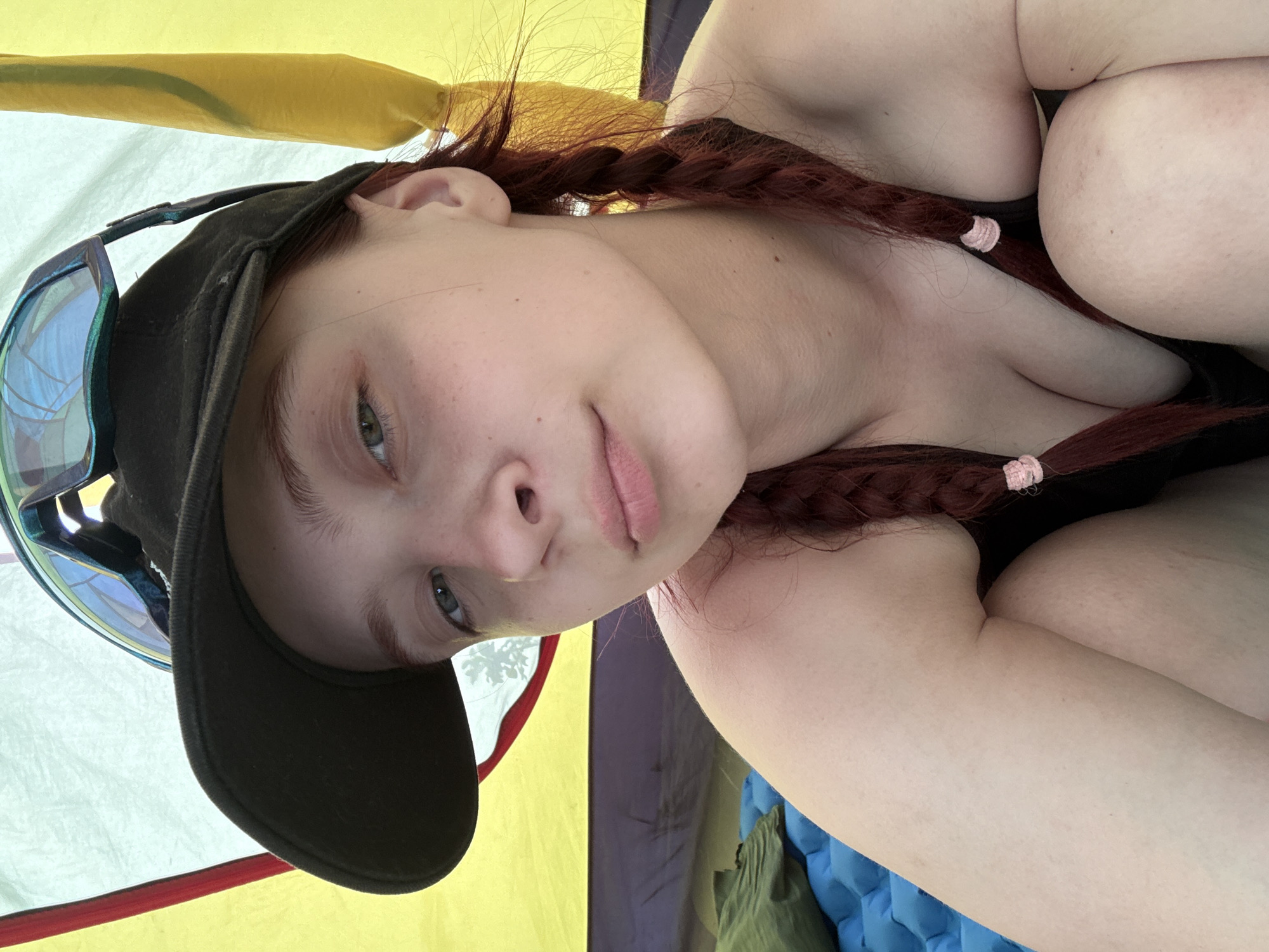 A young woman with fair skin and auburn hair braided into pigtails with pink hair ties poses for a selfie indoors. She is wearing a black cap and a pair of sunglasses, pushed up onto the cap's brim. Her visible attire appears to be black. She is positioned inside what seems to be a tent with panels of yellow, white, and purple fabric. A blue inflatable sleeping pad and a green bag are visible in the lower right corner of the frame. The image's orientation suggests it may have been taken with the phone tilted. There is no information available to confirm the specific location as the city of Khvalynsk, Russia.
