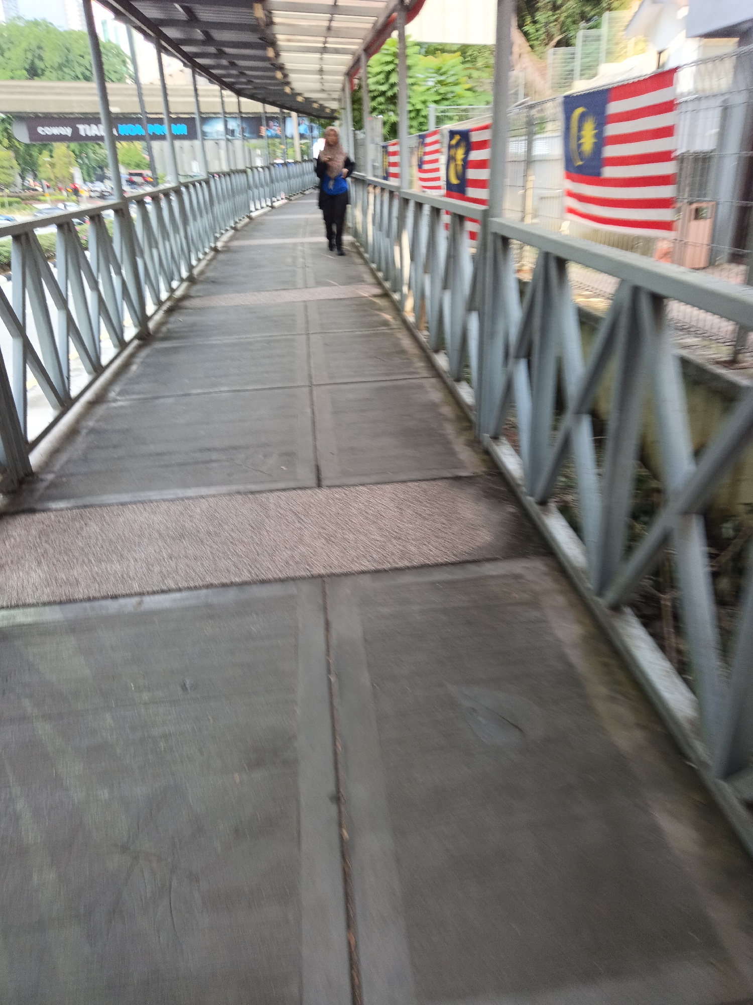 A woman wearing a blue top and a dark headscarf walks along an elevated pedestrian walkway with a grey metal railing on the right side of the frame. The walkway is made of concrete slabs and has a section of darker, rougher material in the middle. Several Malaysian flags are visible along the fence to the right of the walkway. In the background, trees and buildings can be seen. The overall image appears to be taken in Kuala Lumpur, Malaysia, possibly near the KLCC area given the presence of Malaysian flags and the urban environment.