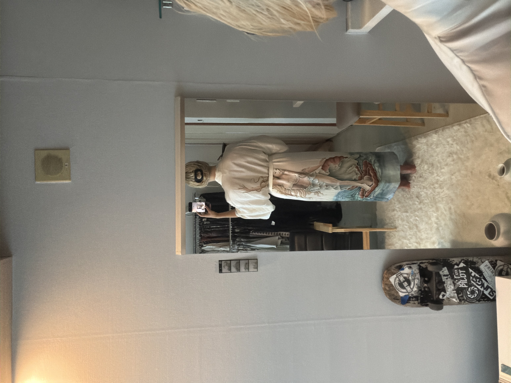 The image appears to be a mirror selfie taken in a room in Seoul, South Korea. A person with blonde hair is partially visible at the top of the image. Their reflection in the mirror shows them wearing a white shirt and patterned pants, holding a phone in their hand, possibly taking the picture. The reflection also shows a closet with clothes hanging inside. On the wall, to the right of the mirror, is a skateboard with stickers. Other objects visible include vases and a white rug. A vent is present on the wall to the left of the mirror.