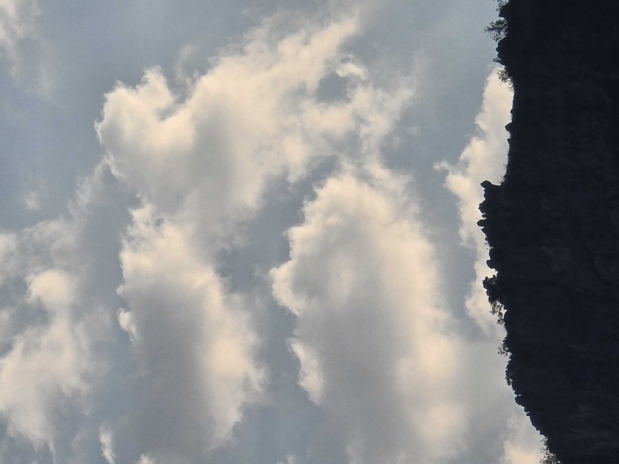 The image shows a silhouetted mountain ridge against a sky filled with dramatic clouds. The mountain ridge is dark and jagged, with some sparse vegetation visible along its crest. The clouds are a mix of bright white and darker gray, suggesting movement and depth. The sky transitions from a light blue at the top to a lighter hue closer to the horizon. There are no people or animals visible in the image. The scene appears to be natural and unpopulated, likely a natural landscape surrounding a city. While the specific location within Podgorica, Montenegro, cannot be precisely determined from the image alone, it depicts a mountainous terrain that is characteristic of areas near the city.