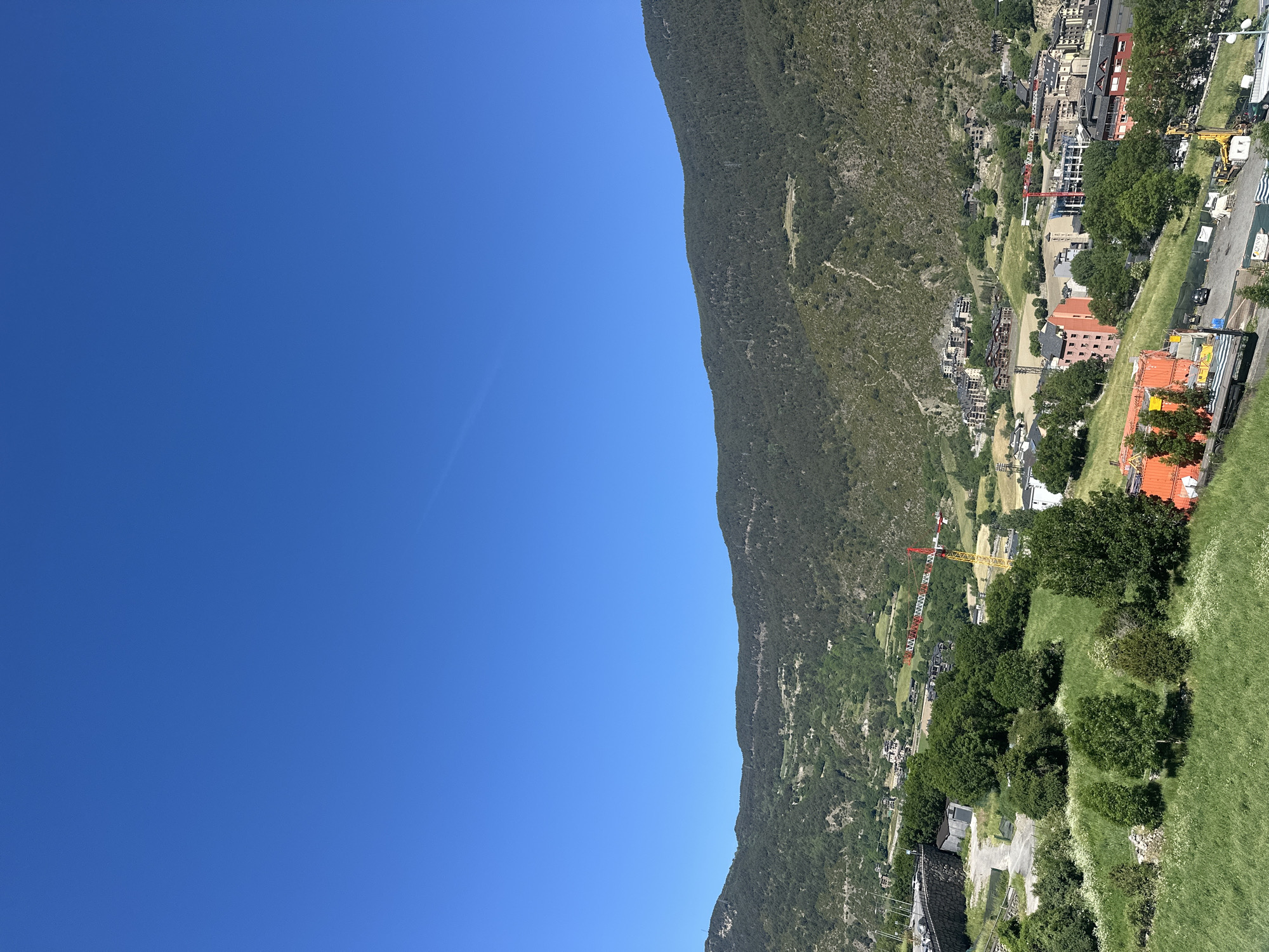 This image provides a high-angle, panoramic view of Encamp, a town situated in a mountainous valley in Andorra, under a clear, bright sky. The original image is rotated, but when viewed correctly, the composition features a vast expanse of intense blue, cloudless sky filling the upper left section.
A prominent, heavily forested mountain slope dominates the right side of the frame, extending from the bottom right towards the top left. Its ridge forms a clean diagonal line separating the sky from the natural and urban landscape. The mountain is covered in dense green vegetation, with some rocky outcrops visible.
The town of Encamp spreads across the lower slopes and valley floor, occupying the mid-ground and foreground. It comprises numerous buildings, including residential and commercial structures, many with traditional sloped roofs, some featuring warm red and orange hues. The town is nestled amongst lush green trees and grassy areas.
Evidence of active development is notable, with several construction cranes visible within the townscape. A large red and white crane stands out in the mid-ground, while a yellow crane is positioned further to the right, indicating ongoing building projects. Roads and various infrastructure elements wind through the town.
The scene depicts a sunny day, likely midday or early afternoon, given the bright illumination and clear shadows. The overall impression is one of a dynamic mountain community coexisting with its dramatic natural environment. No people are clearly visible, and any text on signs is too indistinct to be legible.