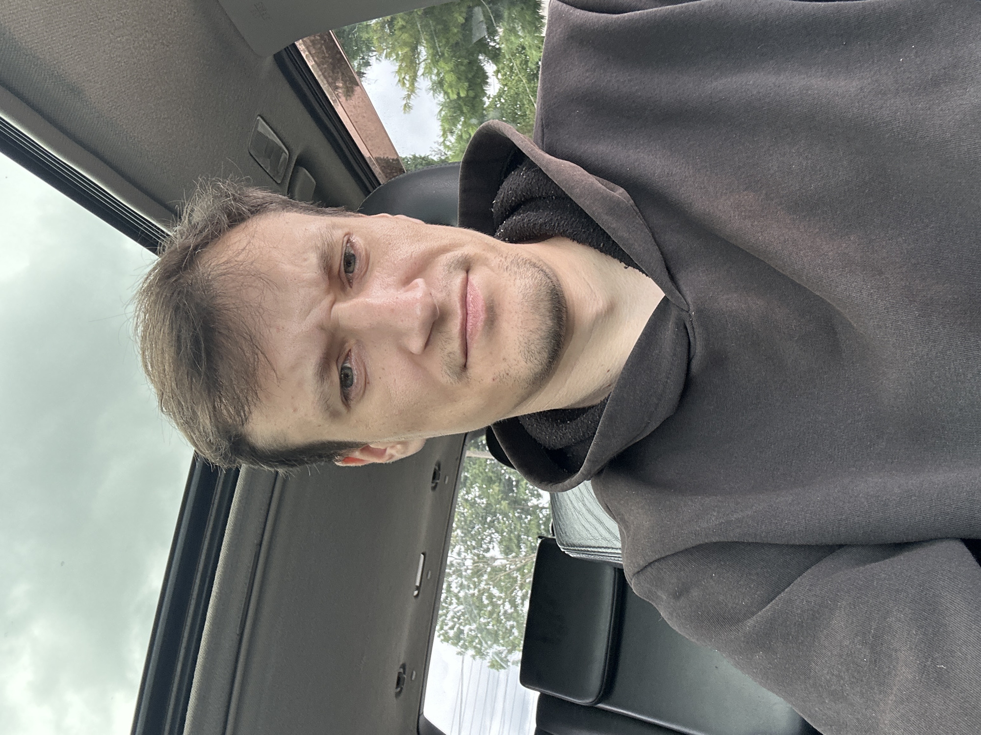 A man with short, brown hair and a skeptical expression sits in the driver's seat of a vehicle. He is wearing a dark brown hooded sweatshirt. The interior of the car is visible, including the black leather seats in the back and a sunroof overhead. Outside the car, trees and an overcast sky are visible. The image is a selfie taken from the man's perspective. The location is Makiv, Ukraine.