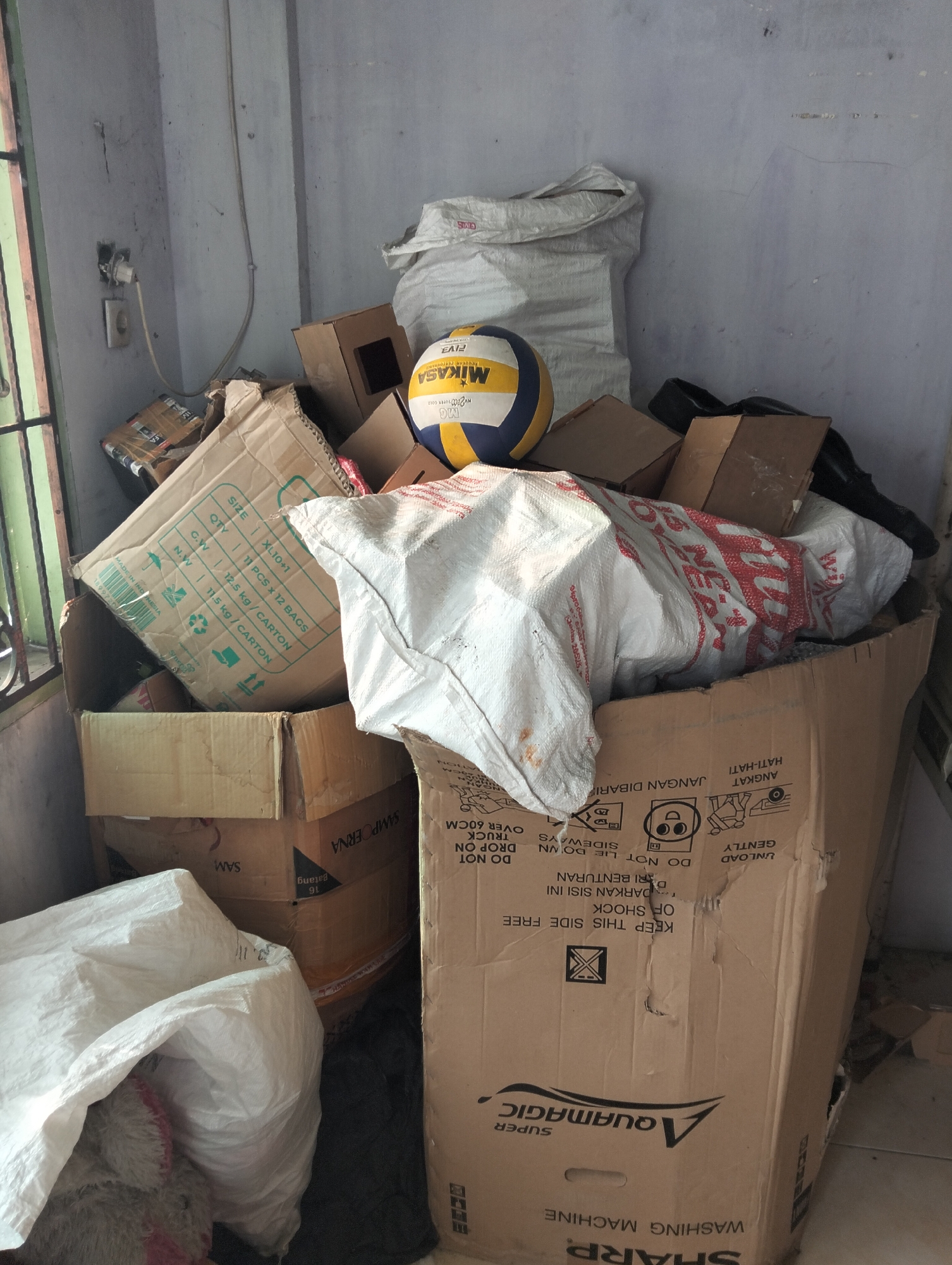 The image depicts a cluttered storage area, likely in a residential or small commercial space, located in the City of Medan, Indonesia. The scene is dominated by a large pile of cardboard boxes, some of which are open, and several large sacks, one of which is white and appears to be made of woven material.
Prominently featured is a volleyball, predominantly yellow and blue, sitting atop a stack of boxes. One large cardboard box on the right side of the image has printed instructions and branding, including the text "Aquamagic SUPER WASHING MACHINE" and "SHARP". Other visible text on boxes indicates "MADE IN INDONESIA" and shipping information such as "SIZE", "QTY", "GW", and "NW". There are also symbols indicating handling instructions, such as "DO NOT LIE DOWN SIDEWAYS" and "GENTLY UNLOAD".
The environment appears to be a room with light-colored, possibly concrete or plaster walls. A window with green-framed bars is visible on the left side. An electrical outlet with a cord plugged into it is mounted on the wall. The overall impression is one of disarray and a temporary holding space for goods. No people are visible in the image. The lighting suggests it is daytime, with some natural light entering from the left.